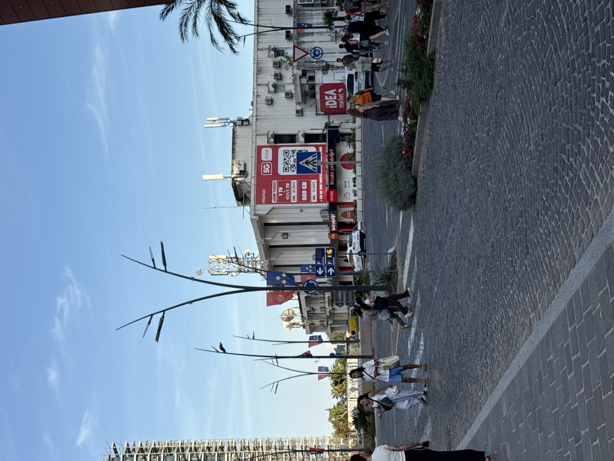 The image shows a street scene in Budva, Montenegro. The perspective is rotated, with the sky at the top left and the street at the bottom right. People are walking along the cobblestone street. Several light poles are visible with decorative branches. Buildings line the street, including one with advertisements for a mobile phone company, Mtel, and a grocery store called Idea Market. The advertisements showcase prices and data plans, mentioning 5G service. Flags from Europe and Montenegro hang from the poles. Traffic signs are visible near a building entrance. Parking signs are displayed above the flags.