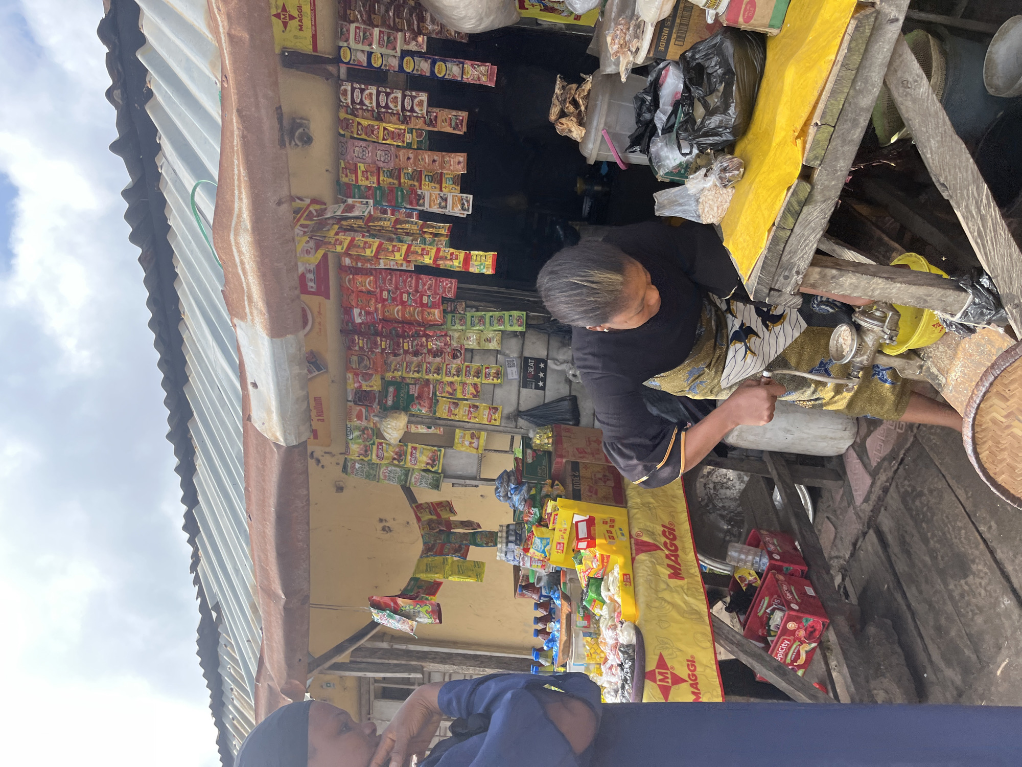 This image shows an outdoor market stall in Jos, Nigeria. The stall is stocked with various food items, including packets of Maggi seasoning and other packaged goods. A woman, likely a vendor, is seen behind the counter, tending to the items. Another person, wearing a blue top, is partially visible in the foreground, appearing to be a customer or passerby. The stall is covered with a corrugated metal roof, and the background reveals a dark interior space filled with more goods. The overall scene suggests a busy marketplace with a focus on food sales. The text "MAGGI" is prominently displayed on yellow banners. The time of day appears to be daytime, with bright natural light illuminating the scene.