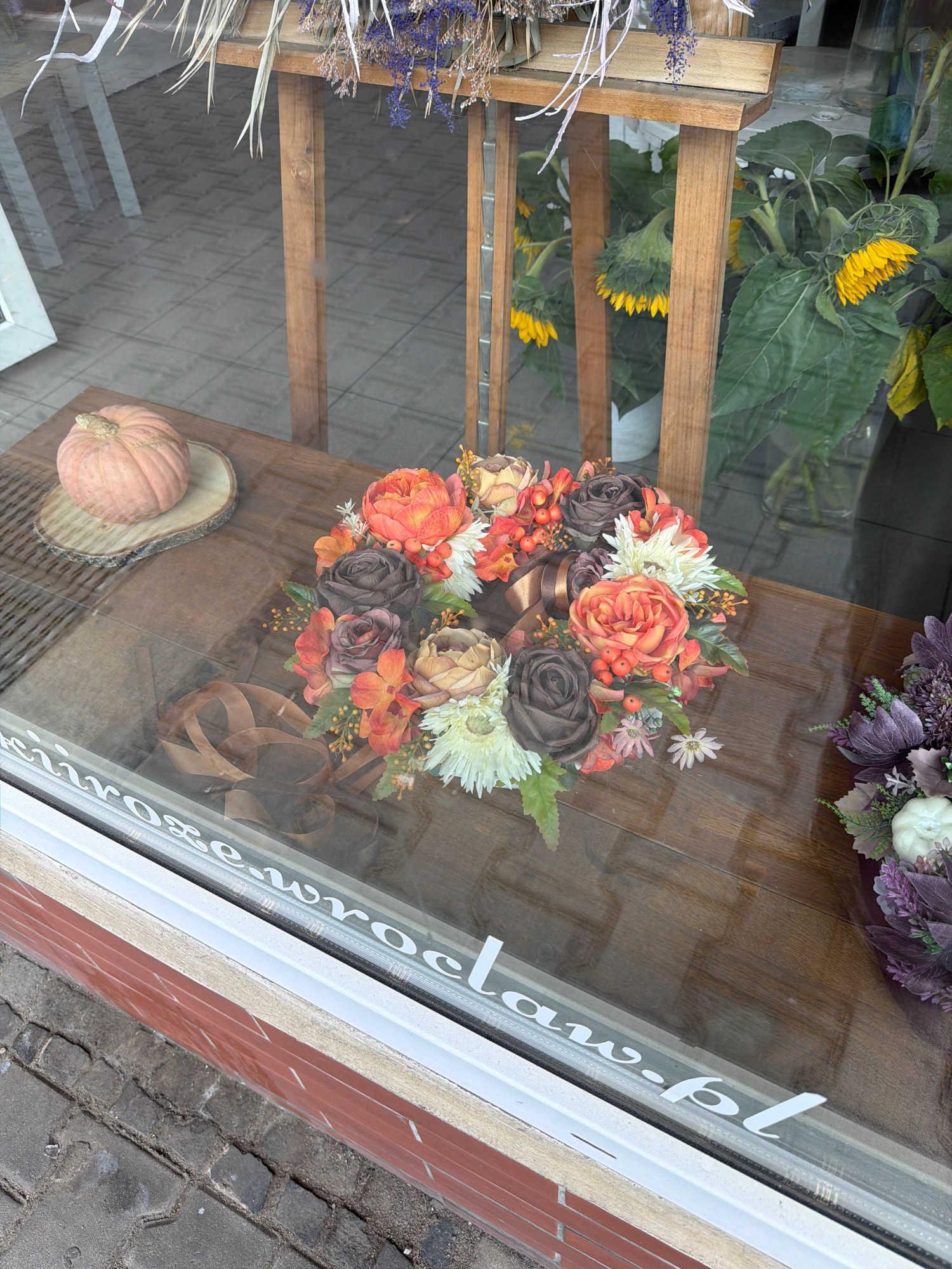A storefront window displays a wooden table with floral arrangements and a small pumpkin. In the foreground, a round floral arrangement features orange, brown, and cream-colored artificial roses with red berries and green leaves. To the left of the arrangement, a small, pale orange pumpkin sits on a wooden slice. A brown ribbon is partially visible beneath the floral arrangement. The window has a white decal with text that reads "czrose.wroclaw.pl", indicating the location of Wrocław, Poland. In the background, through the reflection in the window, a stand with sunflowers is visible.
