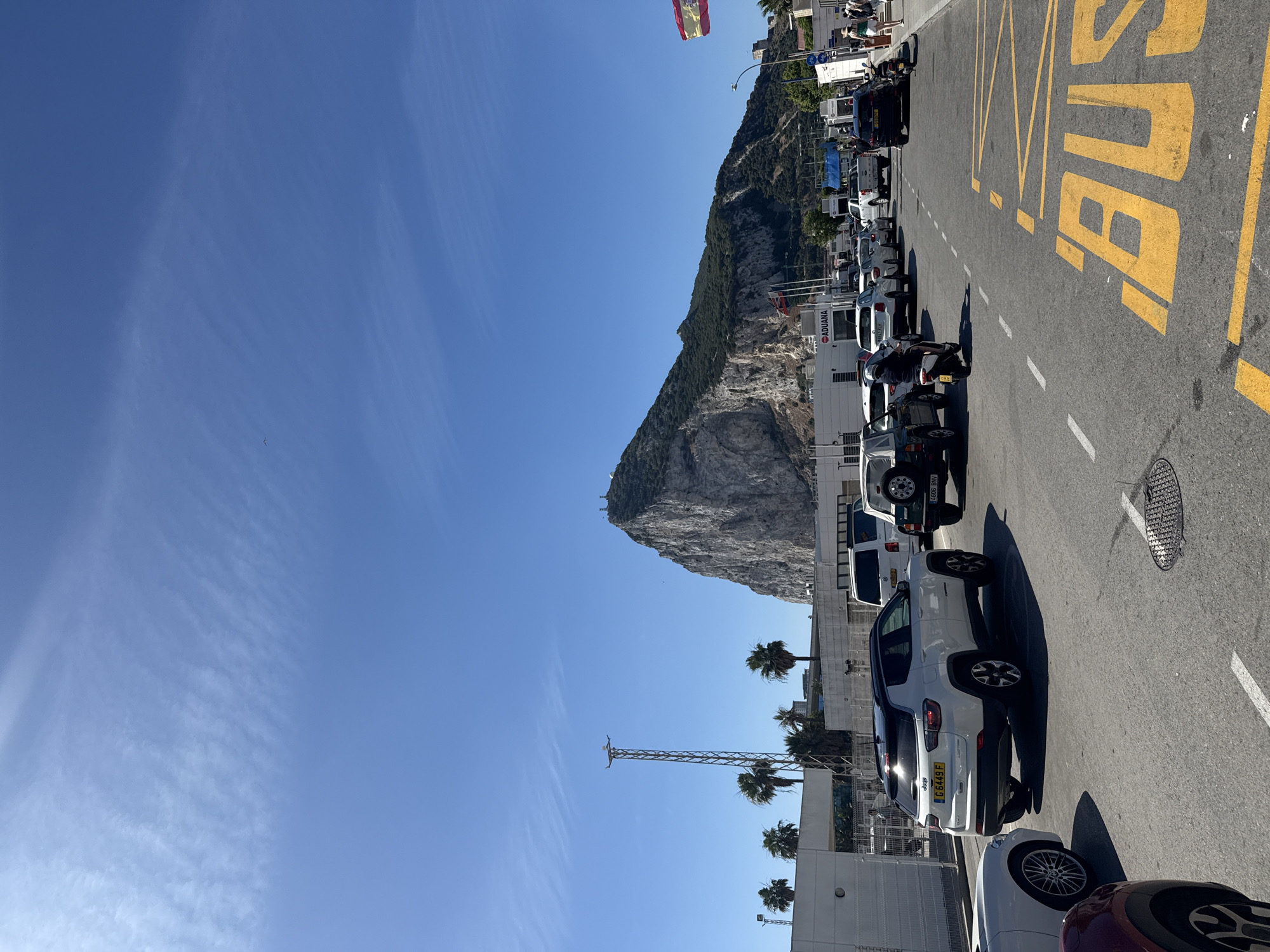 The image shows a sunny day in a border crossing area, likely between Gibraltar and Spain. A prominent rock formation, resembling the Rock of Gibraltar, dominates the background. Several vehicles are parked along the roadside, including cars, a motorcycle, and what appears to be a van or bus. Yellow markings on the asphalt indicate a bus lane. In the distance, a Spanish flag is visible flying from a pole. Palm trees add to the Mediterranean feel of the scene.
On the right side of the image, large yellow lettering on the road clearly spells out "BUS". There is also a sign on a building that reads "ADUANA", which translates to "Customs" in Spanish, further confirming the location as a border crossing. The sky is a clear, bright blue, suggesting fair weather. The time of day appears to be daytime, given the strong sunlight and clear visibility. The overall impression is a busy but orderly scene at an international border.