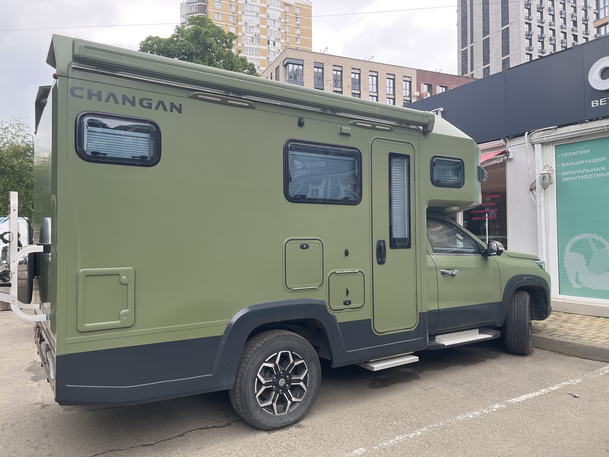 The image shows a green Changan RV parked on asphalt in an urban area. The RV is parked next to a building with a black sign and a vet clinic. The surrounding environment includes apartment buildings and trees. The location is likely Krasnodar, Russia, based on Cyrillic text on a sign in the background. There are no people visible in the image.