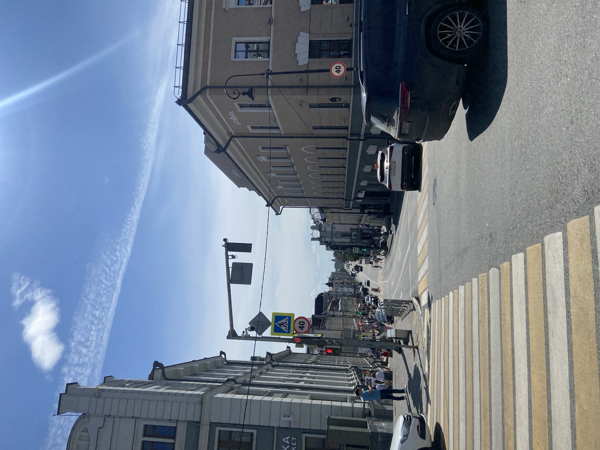 The image displays a sunny daytime street scene in Kazan, Russia, specifically looking downhill on Chernyshevsky Street from its intersection with Kremlevskaya Street. The bright blue sky features a prominent, elongated cirrus cloud streak and a smaller, denser cloud. A strong sunflare emanates from the upper right, casting distinct shadows on the ground.
In the immediate foreground, a broad pedestrian crosswalk with alternating light yellow and white stripes spans the asphalt road. To the left of the crosswalk, a multi-story light grey/white historical building with classical architectural details, including rectangular windows and decorative banding, dominates the left side of the frame. A dark rain gutter is visible on its facade. Partially visible text "KA" and "KET" is present on a sign on this building. On the right side of the street, a multi-story, beige/light brown historical building features numerous windows, ornate facade detailing including a pattern of arches on a lower level, and a decorative black street lamp attached to its corner.
A tall black pole on the left sidewalk supports a red traffic light, a blue square pedestrian crossing sign, a yellow diamond sign, and a circular "40" km/h speed limit sign. Metal railings line the sidewalk along the left side.
Vehicular traffic is present. In the lower right foreground, the rear of a dark-colored SUV, featuring distinctive multi-spoke alloy wheels, is partially visible, casting a large shadow on the road. Behind it, a white SUV with an orange taxi light on its roof and a Russian license plate "Y447HC 116" is visible in the mid-ground, also facing downhill. Multiple other vehicles, including cars and vans, are visible further down the street, either moving or stationary.
Pedestrian activity is evident. On the left sidewalk, a man in a blue t-shirt and light pants stands looking at his phone. Several other pedestrians are visible on the sidewalks on both sides of the street and some are crossing further down, indicating moderate foot traffic. Another circular "40" km/h speed limit sign is visible on a pole in the mid-ground on the right, near the beige building. The street descends noticeably into the distance, lined by historical buildings on both sides.