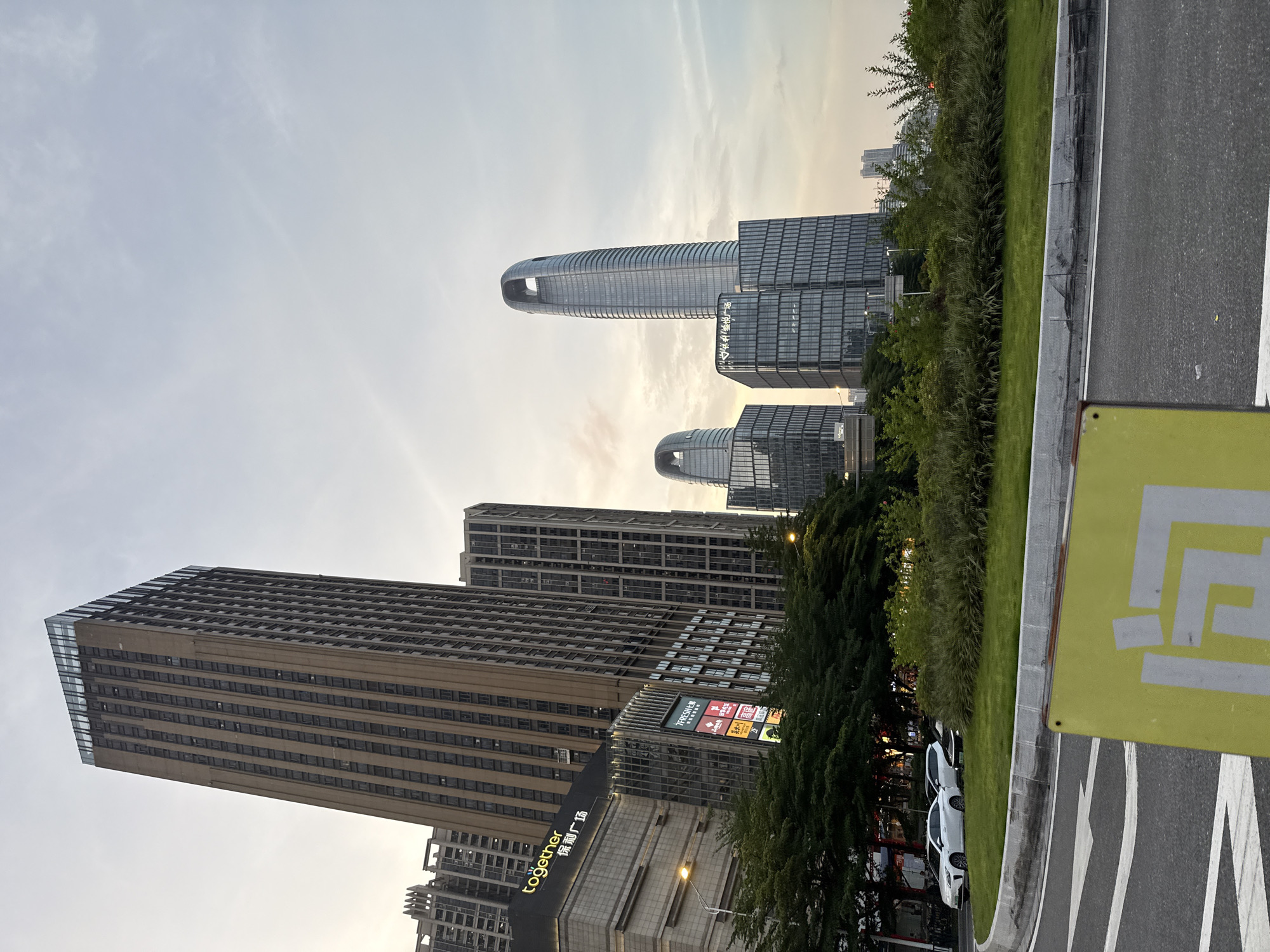 The image depicts an urban landscape in the Haizhu District of China. In the background are modern skyscrapers, including a tall, narrow building with a distinctive ring-shaped top and another with a tiered design. The lower foreground features greenery and trees along a street-level area. Parked cars are visible near the trees. A sign with a pattern in white and light yellow is in the foreground on the right. The sky is light with some cloud formations.