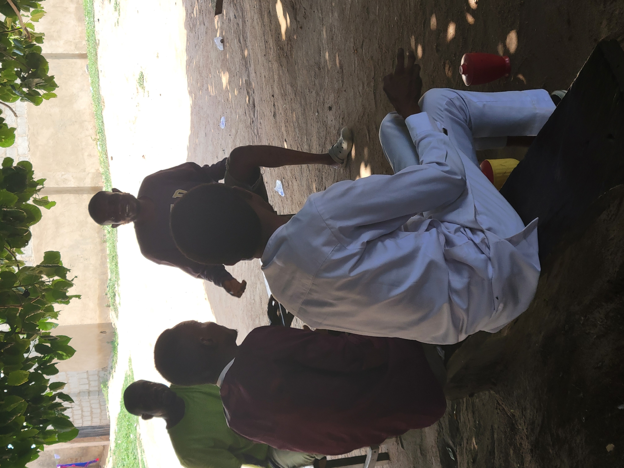 The image depicts a group of four men sitting in an outdoor environment in Madalla, Nigeria. One man is wearing a white shirt and trousers, and he's sitting on what appears to be a wooden plank or bench. To his left, another man is wearing a maroon shirt. The third man is clad in a green shirt. The fourth man is visible in the background, wearing a dark sweatshirt and shorts. The men are sitting together in a shaded area, possibly under a tree. A red plastic container is visible near the man in the white shirt. The ground is mostly dirt with some debris scattered around. A building is visible in the background.