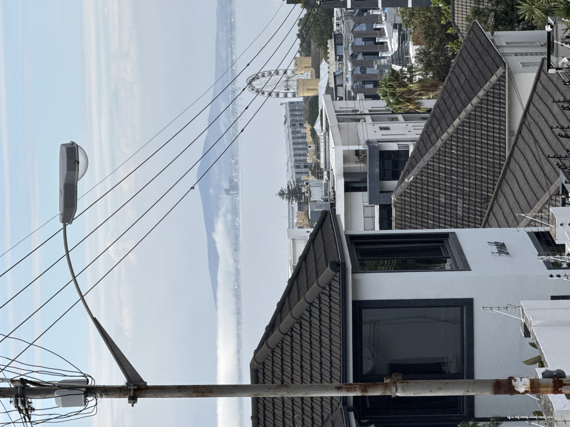 This photograph depicts a streetscape in Cape Town, South Africa, observed from an elevated vantage point. The composition is dominated by rooftops of contemporary residential buildings, characterized by dark-tiled gable roofs and modern architectural styles. A large Ferris wheel is visible in the distance near the coastline, potentially positioned at the Victoria & Albert Waterfront. Overhead, a network of electrical wires crisscrosses the sky, anchored to a utility pole visible to the left of the image. The foreground features a rusted metal pipe or railing, positioned close to the camera, providing a sense of depth and perspective. The sky is overcast, with a pale blue hue, and the distant shoreline appears hazy, suggesting some atmospheric conditions. No people are immediately apparent in the view, but the presence of residential structures implies a residential neighborhood. The overall environment suggests an urban coastal setting.