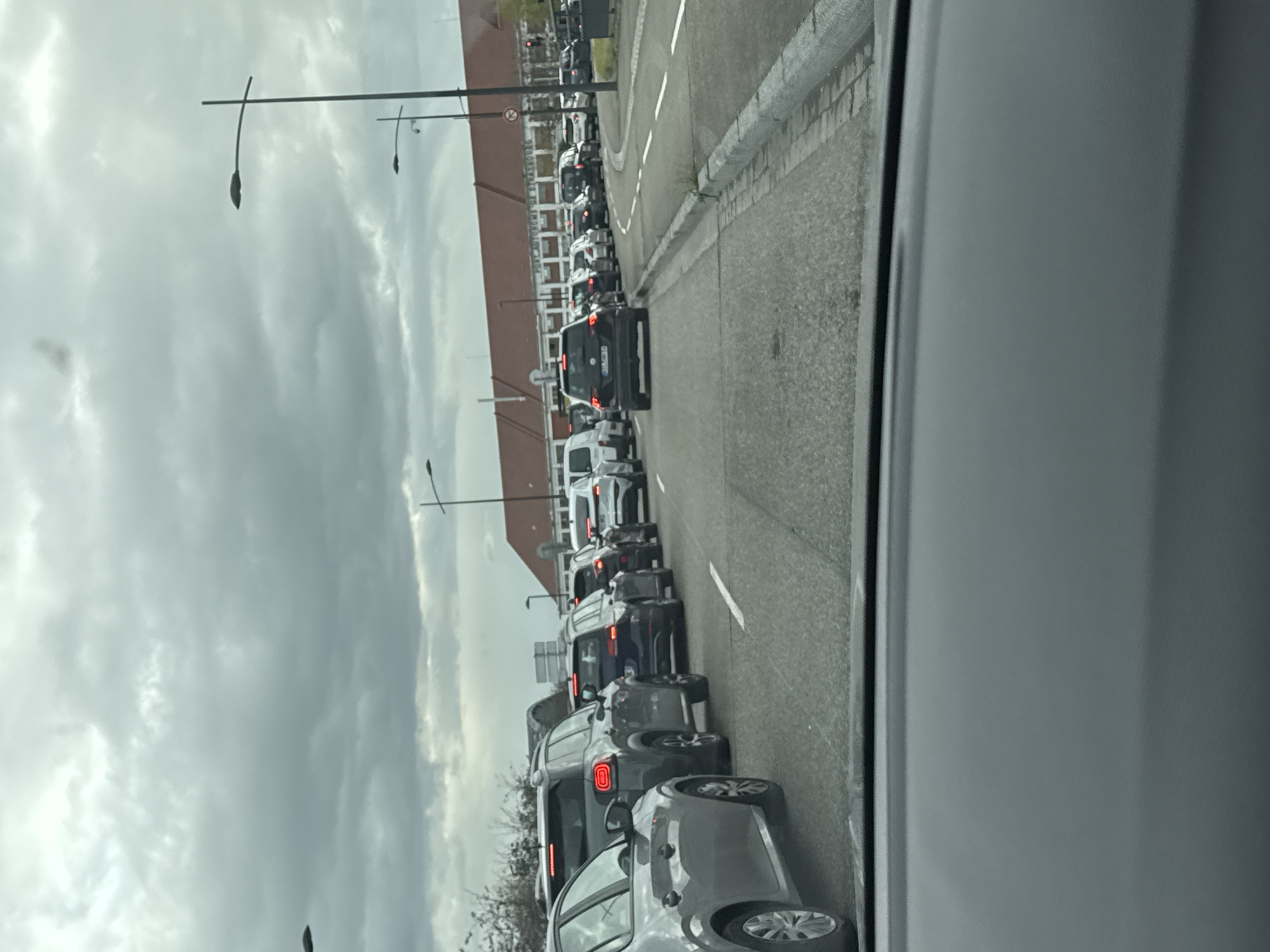The image shows a line of cars stuck in traffic on a road in Strasbourg, France. The cars are lined up along the right side of the road next to a sidewalk. On the other side of the sidewalk is a building with the words "PORT AUTONOME DE STRASBOURG" visible. Street lights are present. The sky is overcast. The image was taken from inside of a car.