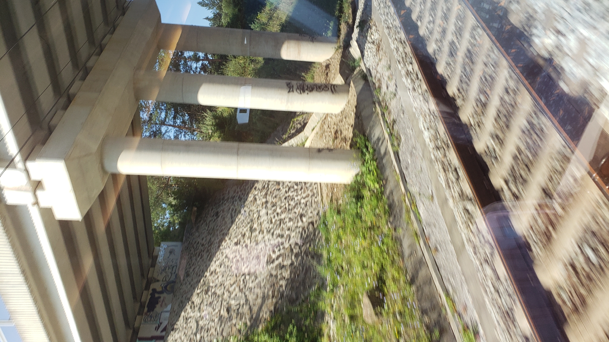 An elevated concrete overpass, supported by thick cylindrical pillars, crosses over a set of train tracks in Horný Hričov, Slovakia. The pillars are marked with spray-painted graffiti. The ground beneath the overpass is covered with loose stones, and a grassy embankment with scattered small flowers runs alongside the tracks. Trees are visible in the background, and the sky is a clear blue. The image appears to have been taken from a moving vehicle, as evidenced by the motion blur on the train tracks and surrounding environment.