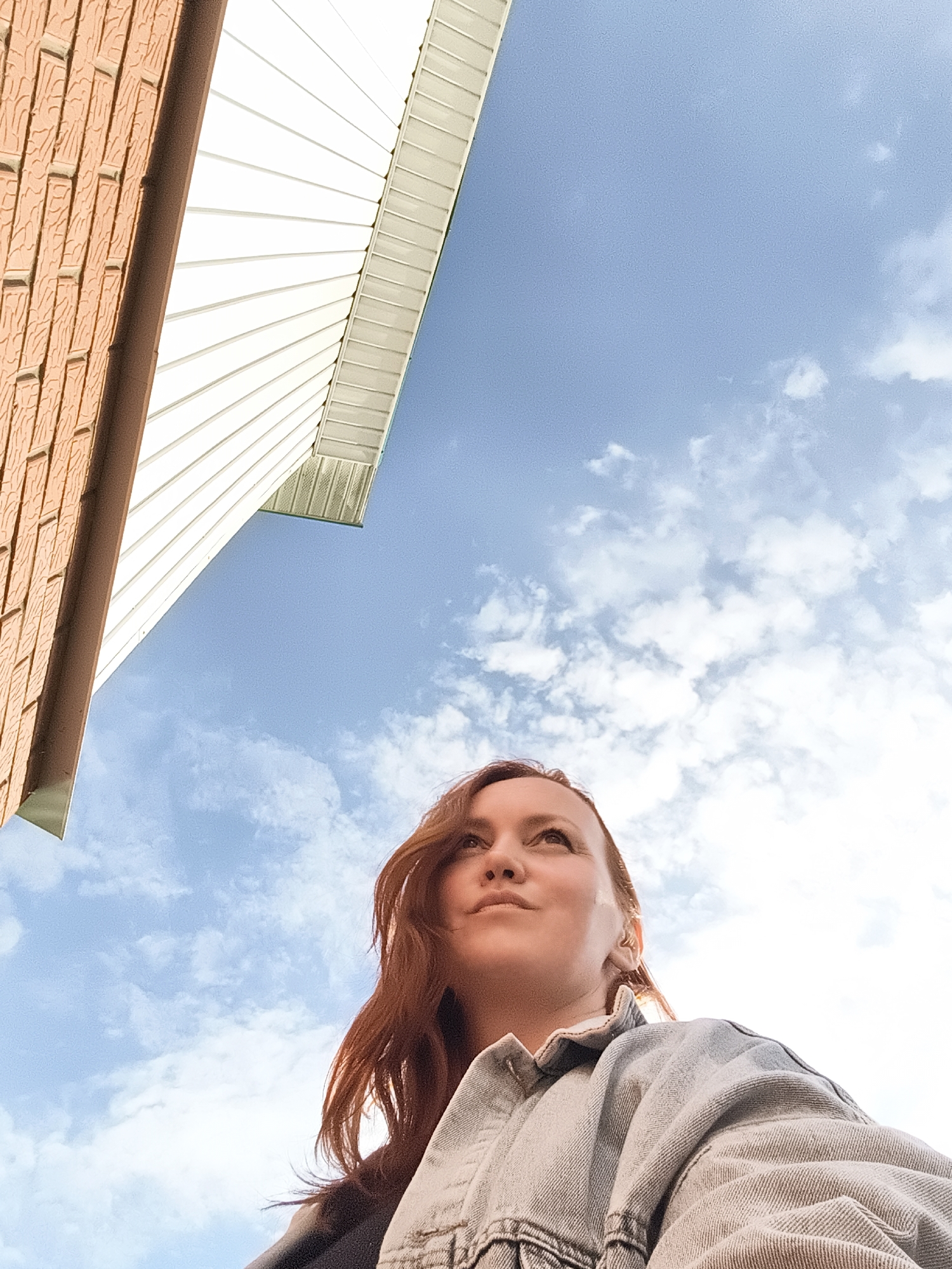 The media file captures a low-angle shot of a woman looking upwards. She has auburn hair and is wearing a denim jacket. The background consists of a bright blue sky with scattered white clouds, suggesting a daytime scene with fair weather. To the left, a section of a building is visible, featuring reddish-brown brickwork and white siding with horizontal lines. The architecture suggests an outdoor urban or suburban setting. The woman's expression appears thoughtful or hopeful. There is no visible text in the image. The overall impression is one of a candid moment, possibly taken outdoors in Chaikovsky, Russia.