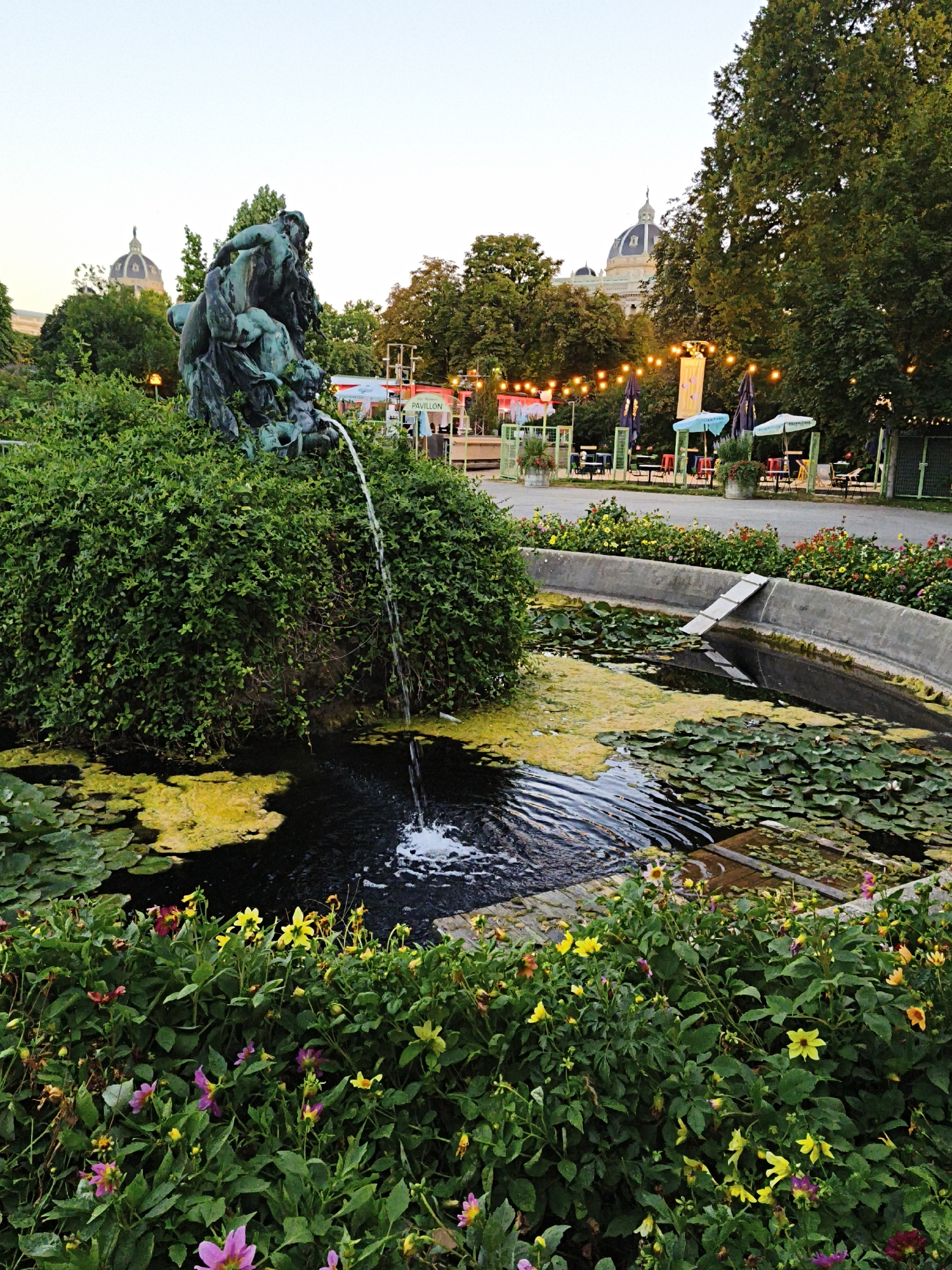 The image depicts a park scene with a fountain in the foreground, featuring a bronze sculpture of several figures from which water cascades into a pond filled with lily pads and algae. The pond is surrounded by lush green foliage and colorful flowers. In the background, there is a paved area with tables and chairs, suggesting an outdoor seating area for a cafe or restaurant. String lights are hung overhead, casting a warm glow. Behind this, trees and buildings with domes can be seen, hinting at a city environment. The architecture of the domes suggests a location in Vienna, Austria, possibly the Volksgarten or Burggarten, known for their fountains and proximity to imperial buildings. The lighting indicates it is either early morning or late evening. There are no people clearly visible in the scene.