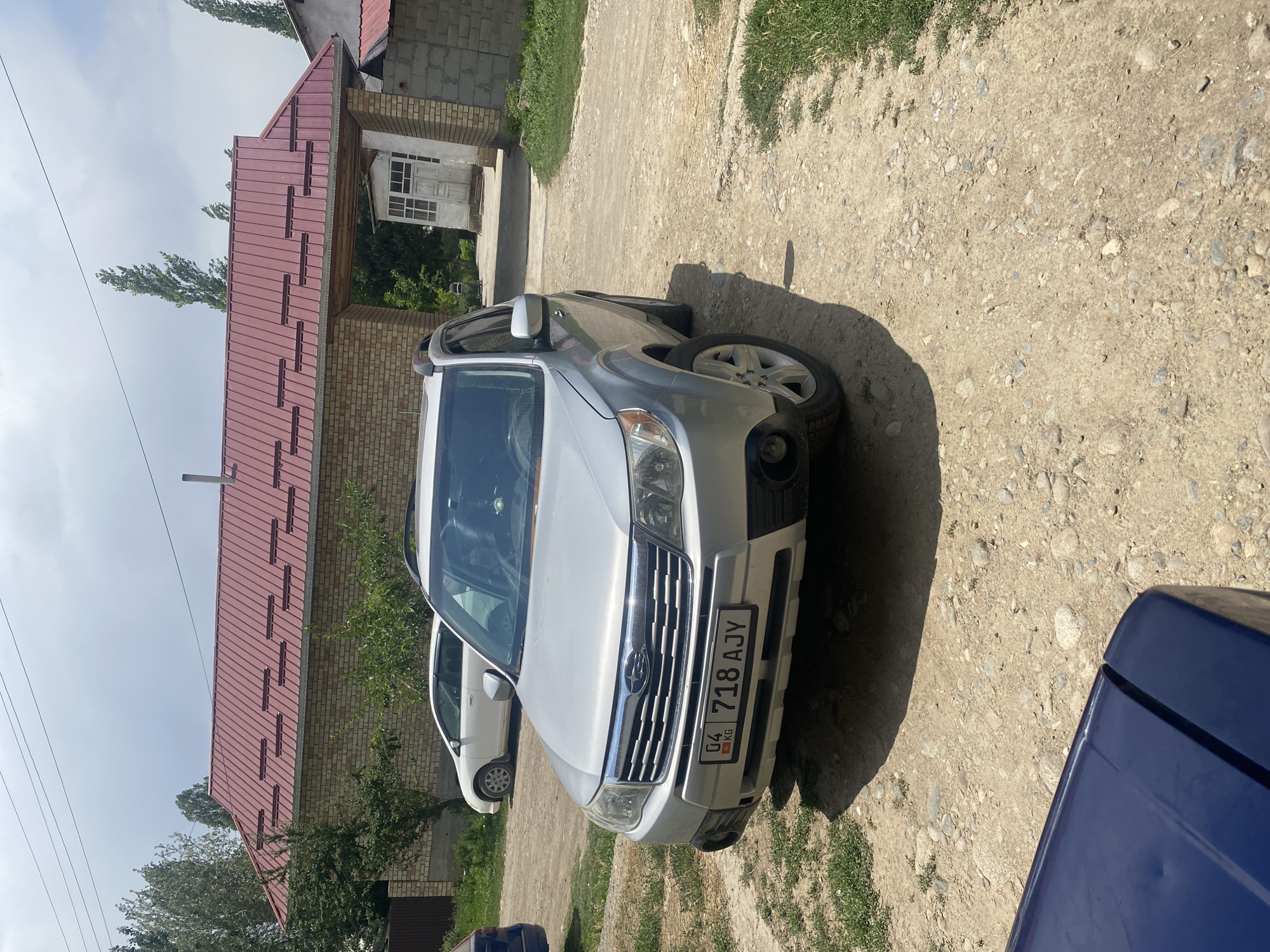 A silver Subaru Forester SUV is parked on a dirt road in front of a brick building with a red metal roof. The license plate of the SUV reads "04 KG 718 AJY". A white car is parked to the left of the SUV. Trees and green grass are visible in the background. The sky is cloudy. The scene appears to be a residential area.