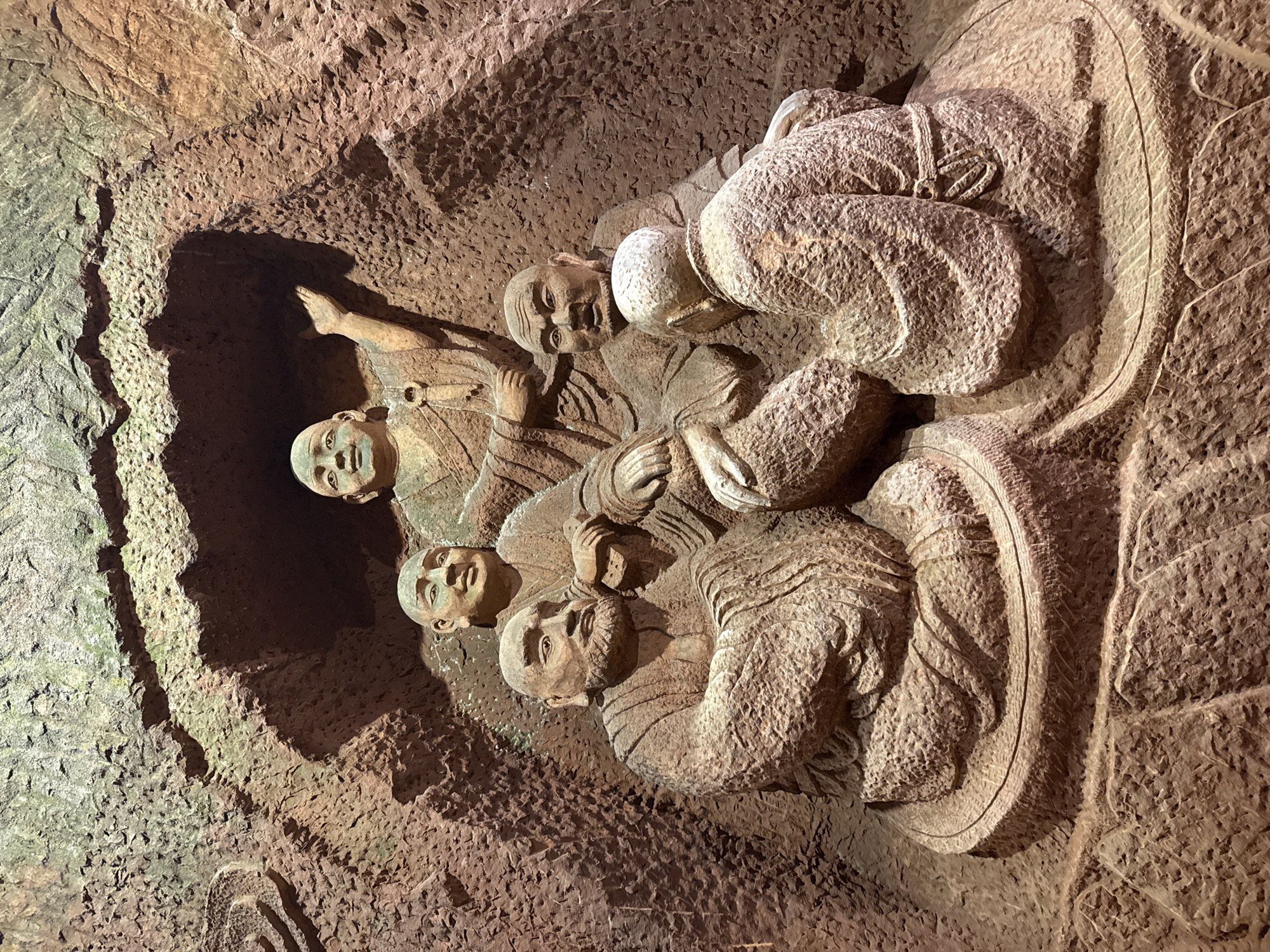 This image depicts a stone carving of five figures in an unknown city and country. The carving is embedded within a roughly hewn stone surface that forms a wall or a natural cave-like environment. The figures, dressed in what appear to be simple robes, are grouped closely together within a carved, roughly circular or oval frame. Their facial expressions seem jovial or contemplative. The figures are of East Asian descent. The carving's texture is coarse and detailed.