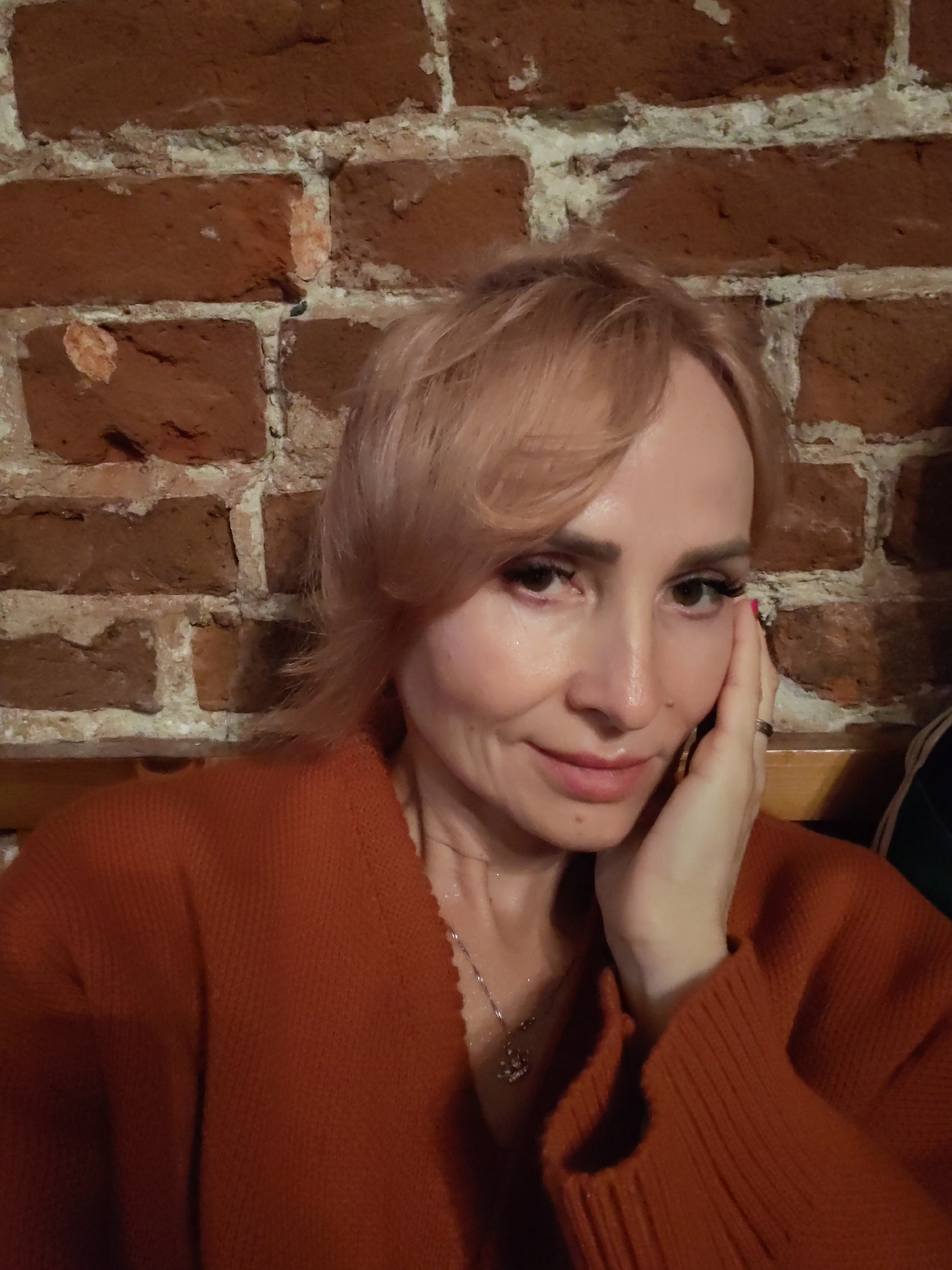 The image is a medium shot of a woman with blonde hair and an orange sweater. She is resting her right hand on her cheek, and her left hand is visible on her chest. The background is a brick wall. The lighting appears to be indoor, and the mood seems relaxed. Based on the provided location context, this photo was likely taken in Nizhny Novgorod, Russia. There is no visible text in the image.