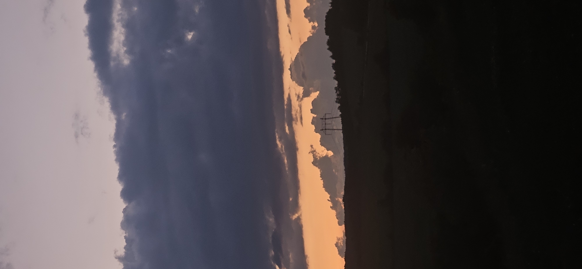 The image depicts a twilight landscape in Gutsal, Bulgaria. The sky is divided into distinct layers: the upper two-thirds are dominated by dark, heavy stratocumulus clouds in shades of deep grey and muted purple. Below these, a prominent horizontal band of vibrant orange and lighter pink light indicates a sunset. This orange band illuminates the undersides of smaller, irregularly shaped clouds, creating a contrast against the darker clouds above. On the distant horizon, a large metal electricity pylon, consistent with a high-voltage transmission tower, is silhouetted against the orange sky, positioned slightly right of the vertical center. The undulating horizon line consists of silhouetted terrain, likely hills or mountains, topped with dark, indistinct vegetation, possibly trees. The entire lower third of the image is rendered in deep black silhouette, representing the immediate foreground and obscuring any ground details. No people or animals are visible.