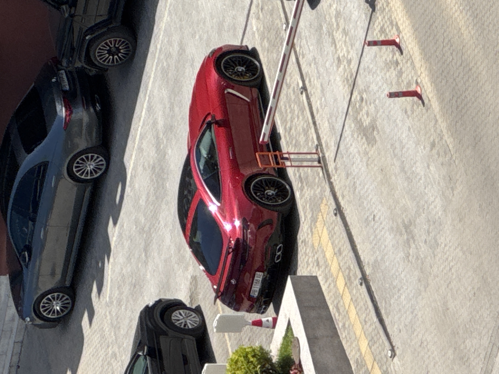 Captured from an elevated perspective, the image depicts a parking area in Plovdiv, Bulgaria. A red sports car is prominently positioned next to a barrier and close to traffic cones. To the left, a dark-colored passenger car is parked, and another car is partially visible. The area appears to be a paved lot with lane markings. Vegetation is present.