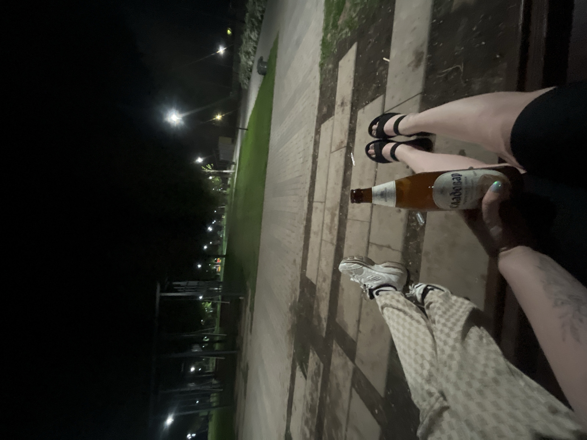 At night, in what is likely a park in Zav'yalovo, Russia, two individuals are sitting on a bench. One person is wearing black sandals and black shorts, while the other has on sneakers and checkered pants. One person is holding a beer bottle labeled "Clausthaler". The background shows a park area with trees and street lights.