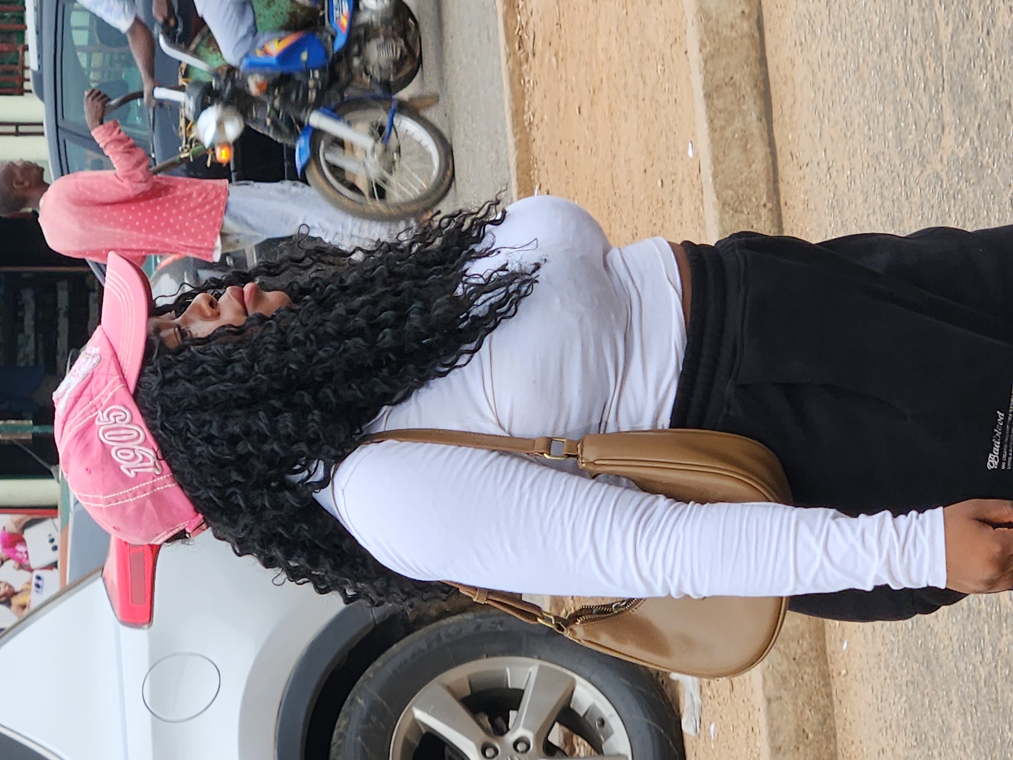 A medium shot shows a young Black woman in profile, wearing a pink baseball cap with "1905" written on it, a white long-sleeved crop top, and black sweatpants. She has long, curly black hair and a tan shoulder bag. To her left is the rear of a silver SUV, and behind her, a man in a pink, polka-dotted shirt stands near a black SUV, with a blue motorcycle partially visible in the background. The scene appears to be on a street with a dirt and asphalt surface, bordered by a concrete curb.