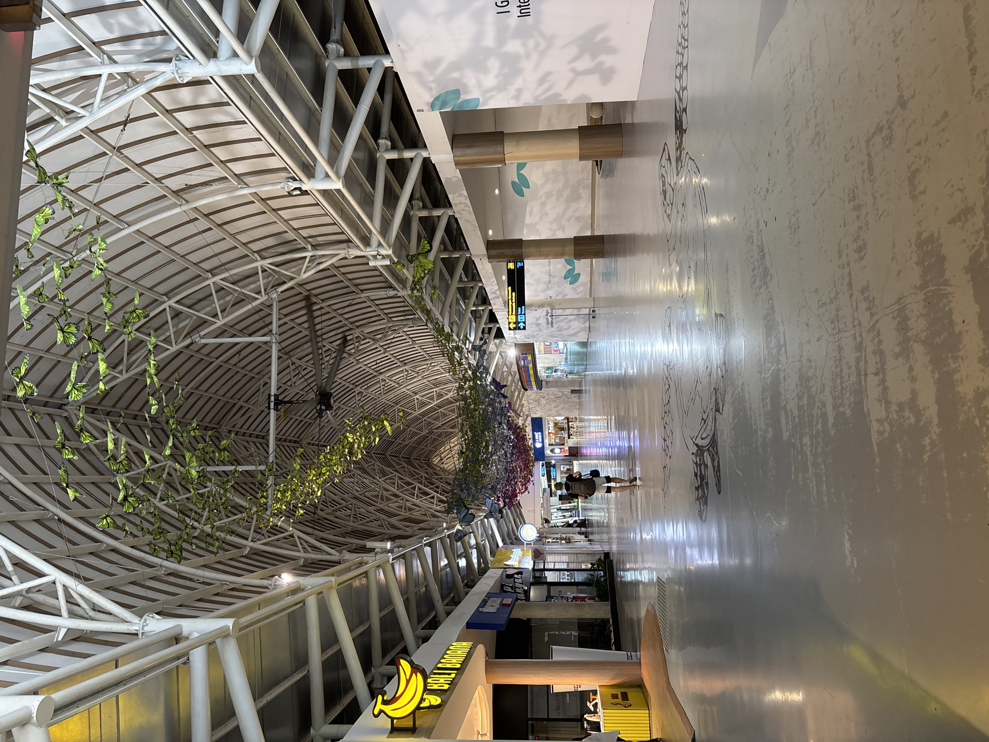 This image shows the interior of the I Gusti Ngurah Rai International Airport in Kuta, Bali, Indonesia. The vaulted ceiling is constructed of white steel beams and decorated with hanging butterfly and floral arrangements. A single person walks down the center of the hallway. Retail spaces line the bottom of the hallway. The floor is light-colored with sketched decoration. Signage is partially visible.