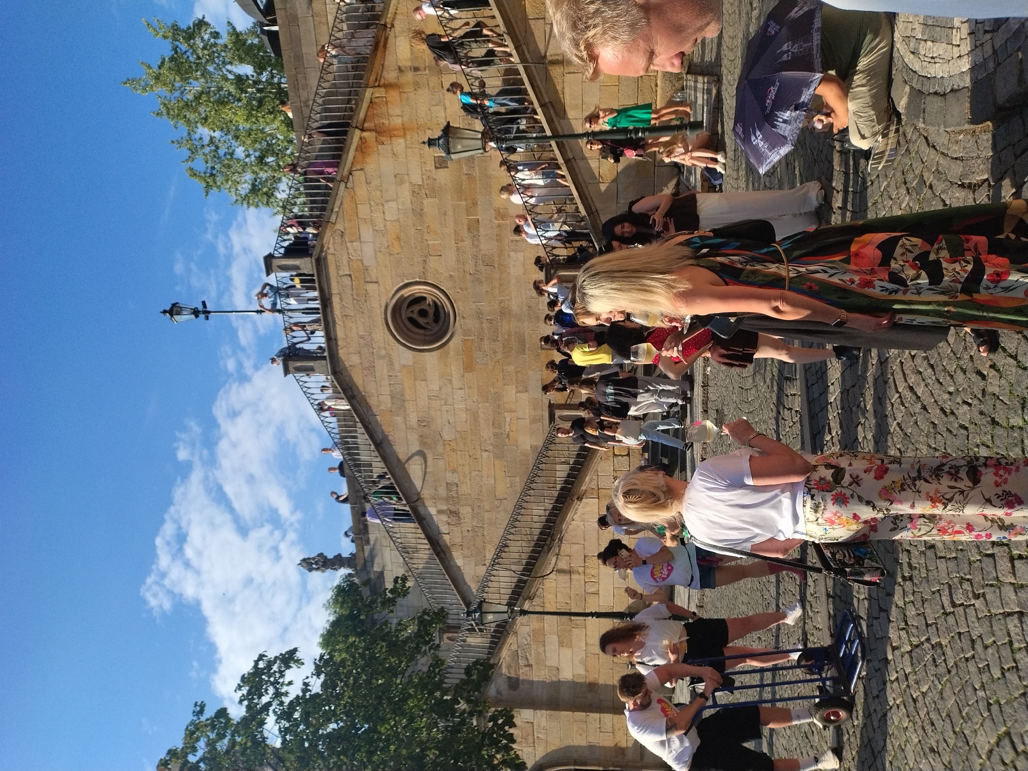 A street-level view captures a bustling outdoor scene in Prague, Czechia, with a large stone staircase dominating the background and a cobblestone street in the foreground. The staircase, made of light-colored stone blocks, features numerous steps and is lined with metal railings. Several people are ascending and descending the stairs, some pausing to look around or take photos.
On the cobblestone street, a diverse group of people are gathered. In the foreground, a woman in a white t-shirt and a long, colorful floral skirt stands facing away from the camera, holding a glass of white wine. To her left, a man in a white t-shirt and black shorts is pushing a blue trolley. Other individuals are engaged in conversations, holding drinks, or observing the surroundings.
The environment is illuminated by bright sunlight, casting shadows on the cobblestones. A street lamp with an ornate design stands near the staircase. Trees with green foliage are visible in the upper left portion of the image. In the lower right corner, a person is seated on the ground under a dark umbrella, appearing to be selling or offering something from a basket. The sky above is blue with scattered white clouds.