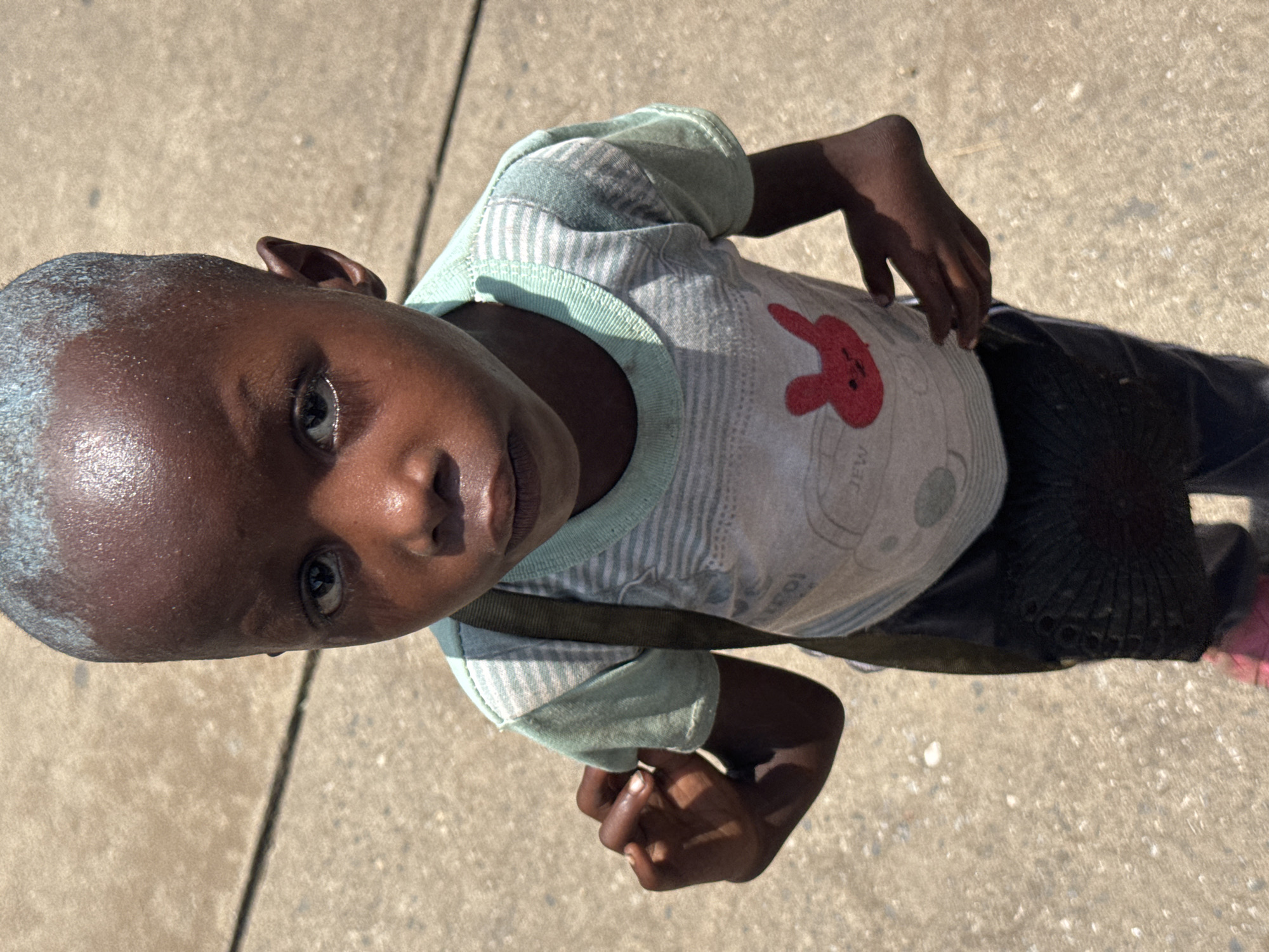 A young Black child is depicted in a high-angle, top-down shot, looking directly into the camera. The child has very short, close-cropped hair with a lighter, possibly dusty or painted, residue visible on the scalp. A slight sheen on the forehead and cheeks suggests perspiration.
The child is wearing a light-colored t-shirt with alternating thin white and light green stripes. The collar and sleeve cuffs are light green. A prominent red graphic of a rabbit's head is on the front of the t-shirt, positioned towards the child's left. Faint, unreadable text appears on the striped pattern of the shirt and below the rabbit graphic. A dark strap, possibly from a bag or satchel, crosses diagonally over the child's chest from the left shoulder towards the right hip. Dark trousers or pants are visible below the t-shirt, and a dark, textured object, possibly a crumpled bag or fabric, is tucked on the child's right side, partially obscured by the strap.
The child's hands are loosely clasped or resting in front, partially visible. The setting is an outdoor, brightly lit environment, indicated by strong highlights and shadows. The child stands on a light-colored, rough-textured ground surface, likely concrete or asphalt, with visible seams and light speckling. The perspective creates an intimate and direct engagement with the viewer. The time of day appears to be sunny, likely midday or early afternoon, consistent with a warm climate like Onitsha, Nigeria. The child's expression is direct and observant.