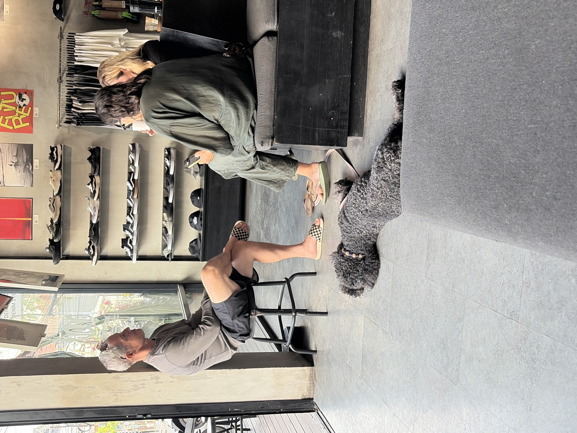 In North Kuta, Indonesia, a casual scene unfolds inside a retail establishment with concrete floors and grey walls. A grey-haired man with sunglasses perched on his head sits on a black metal stool, his legs crossed, wearing a long-sleeved grey shirt and black shorts. He is looking to his left. Beside him, a black poodle lies on the floor, its body stretched out.
Further into the store, two women are seated on a dark grey sofa. One woman, facing away from the camera, wears a dark olive green oversized garment and holds a phone in her right hand. The other woman, with blonde hair and wearing a black top, is partially visible behind her.
The background reveals shelves filled with footwear, predominantly sneakers, and a rack of clothing, including t-shirts. Posters are displayed on the wall. A large window offers a view of the outdoors.