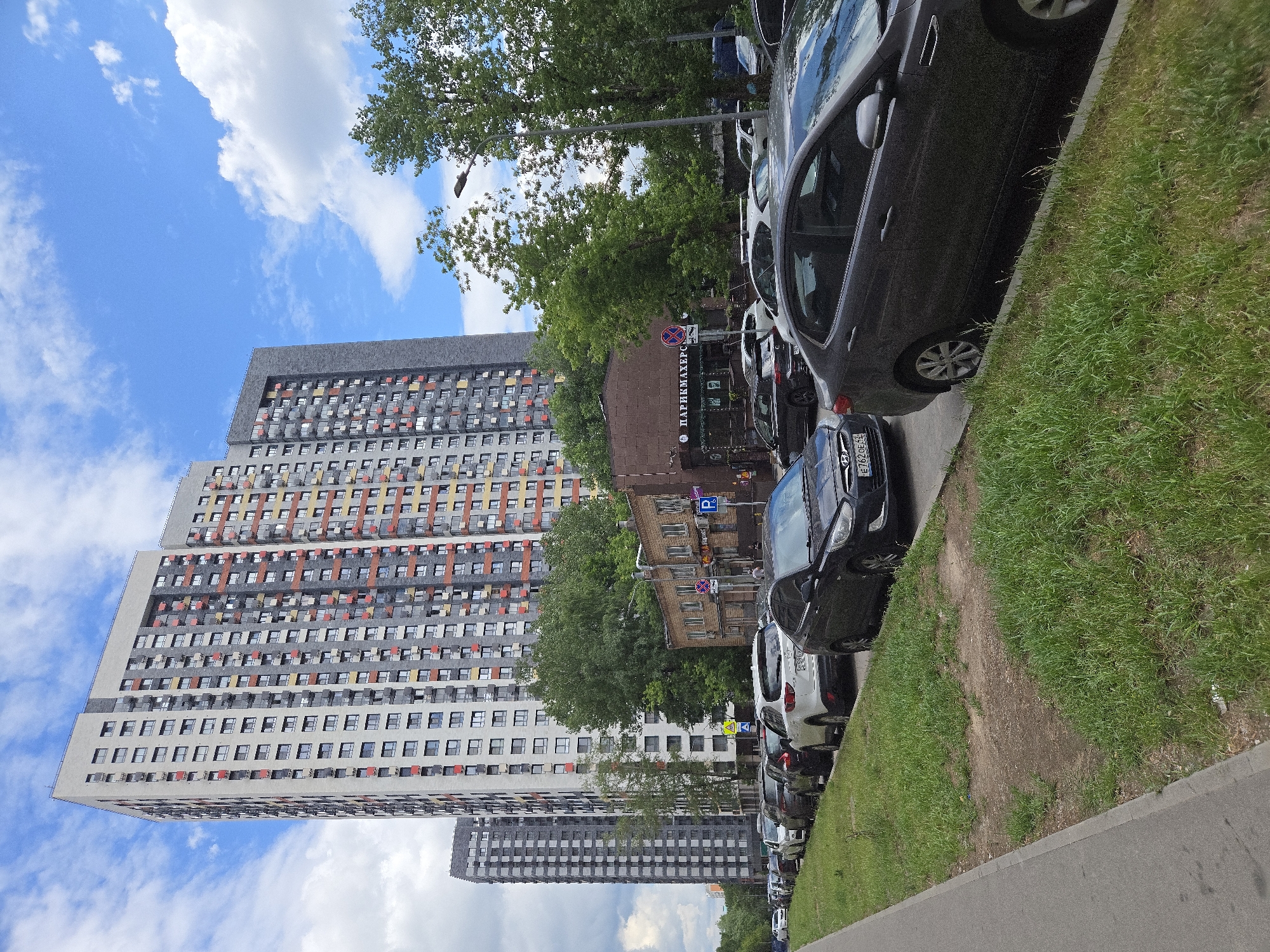 The image depicts a street scene in Moscow, Russia, with a row of parked cars lining a grassy area next to a sidewalk. In the background, several modern, high-rise apartment buildings are visible, characterized by their colorful facades and numerous windows. Lush green trees are interspersed among the buildings, and the sky above is a bright blue with scattered white clouds, suggesting a pleasant daytime weather.
The cars are parked side-by-side along the curb, with their reflections visible on the street. The composition is dominated by the architecture and the vehicles, with the natural elements of grass and trees providing a contrast. A street lamp is visible amongst the trees.
There is some visible text on a sign in the middle of the frame, which appears to be in Russian. It reads "ПАРКМАХЕРСКАЯ" (PARIKMAHERSKAYA), which translates to "barbershop." License plates on the cars are also visible, with some alphanumeric characters readable, such as "E762OE55". The overall impression is that of an urban residential area on a clear day.