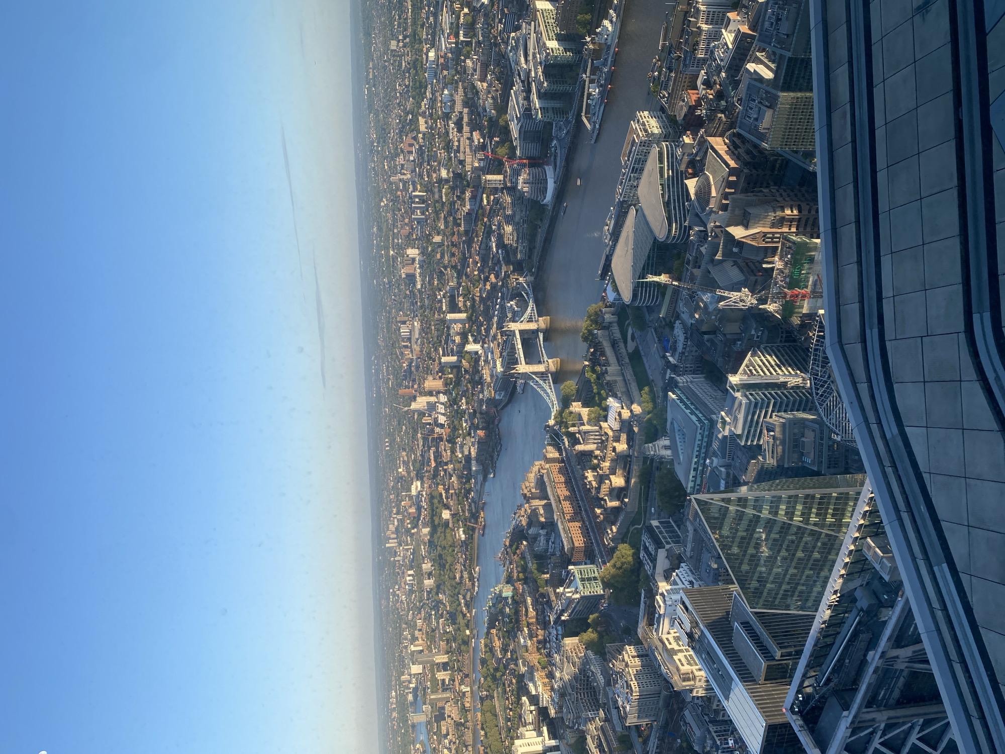 This media file presents a wide aerial panoramic view of London, United Kingdom, captured from a very high vantage point. The scene is dominated by a sprawling urban landscape under a clear, pale blue sky, suggesting late afternoon or early evening due to the soft, warm light and shadows.
In the foreground, on the right side, a substantial portion of a modern, multi-storied skyscraper with a curved, tiled facade is visible, indicating the likely source of the photograph. This structure frames the expansive cityscape beyond.
The majestic River Thames flows prominently through the middle ground, snaking from the left side of the frame towards the right. Numerous iconic landmarks and structures line its banks and cross its expanse. The distinct gothic towers and suspension structure of Tower Bridge are clearly identifiable, positioned in the middle-left, confirming the location. Further along the river, other bridges and various vessels can be seen.
The cityscape extends densely into the background, showcasing a mix of historical and contemporary architecture, including numerous glass-fronted high-rises and smaller, more traditional buildings. Patches of green spaces and trees are interspersed within the urban fabric. Several construction cranes are visible, indicating ongoing development.
A notable detail, upon closer inspection, is text visible on a building or construction site banner. The words "FIFTY TWO" are clearly discernible, likely referencing "FiftyTwo Lime Street," also known as The Scalpel, a distinctive skyscraper in the City of London.
The weather appears clear, though a light atmospheric haze softens the details of the distant city towards the horizon. No specific human activities or individuals are discernible due to the vast distance and elevation. The overall impression is one of a bustling, vast metropolis, seen from an elevated, serene perspective.