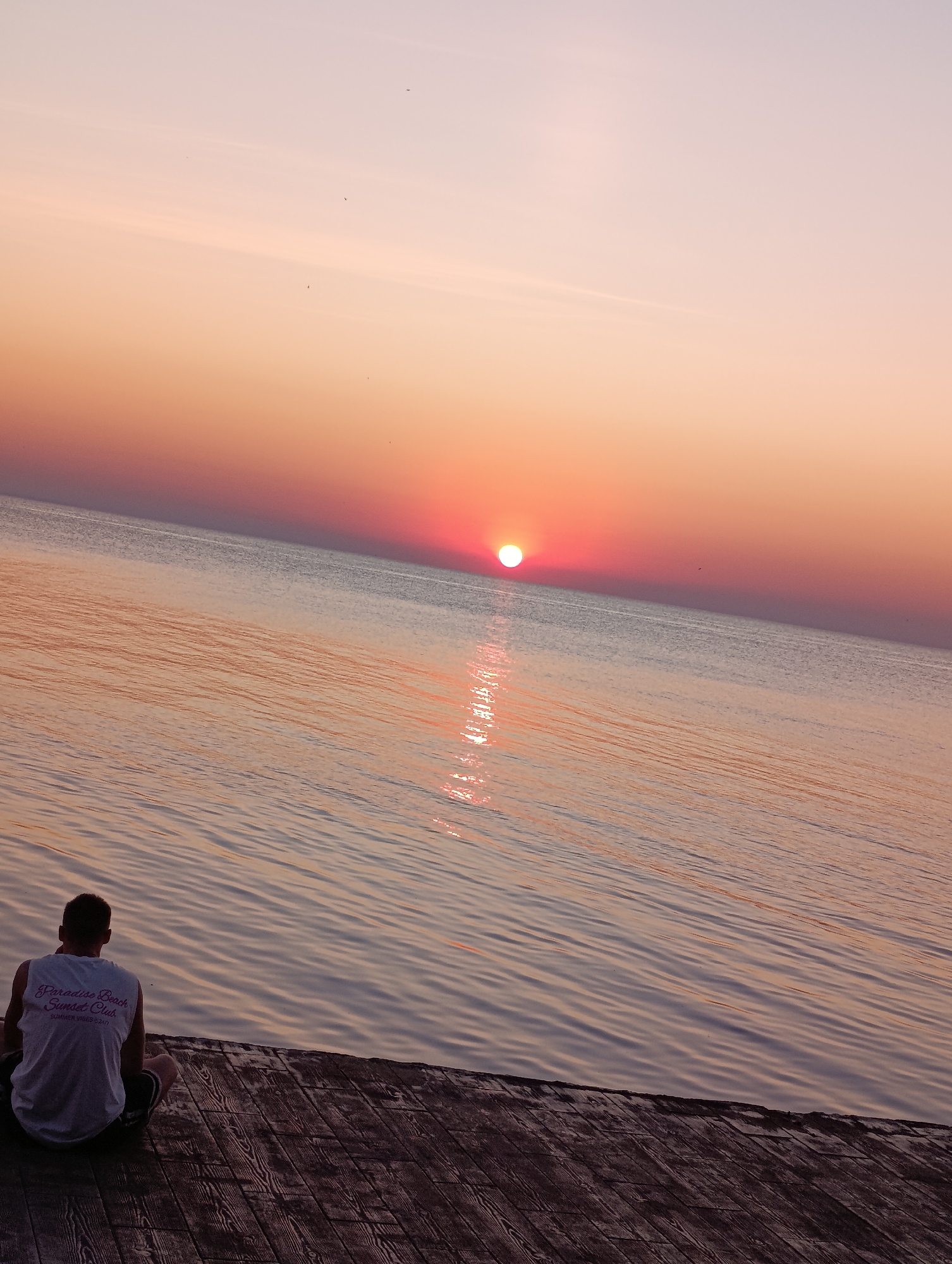 A person sits on a wooden pier, facing a body of water with a sunset in the background. The sky is a gradient of pink and orange, with the sun appearing as a bright orange orb on the horizon. The water is calm, with ripples reflecting the colors of the sky. The pier is made of weathered wooden planks. The person is wearing a white tank top with text printed on the back and dark shorts. The scene appears to be a serene moment of contemplation by the sea. The location is the Odesa embankment, Ukraine.