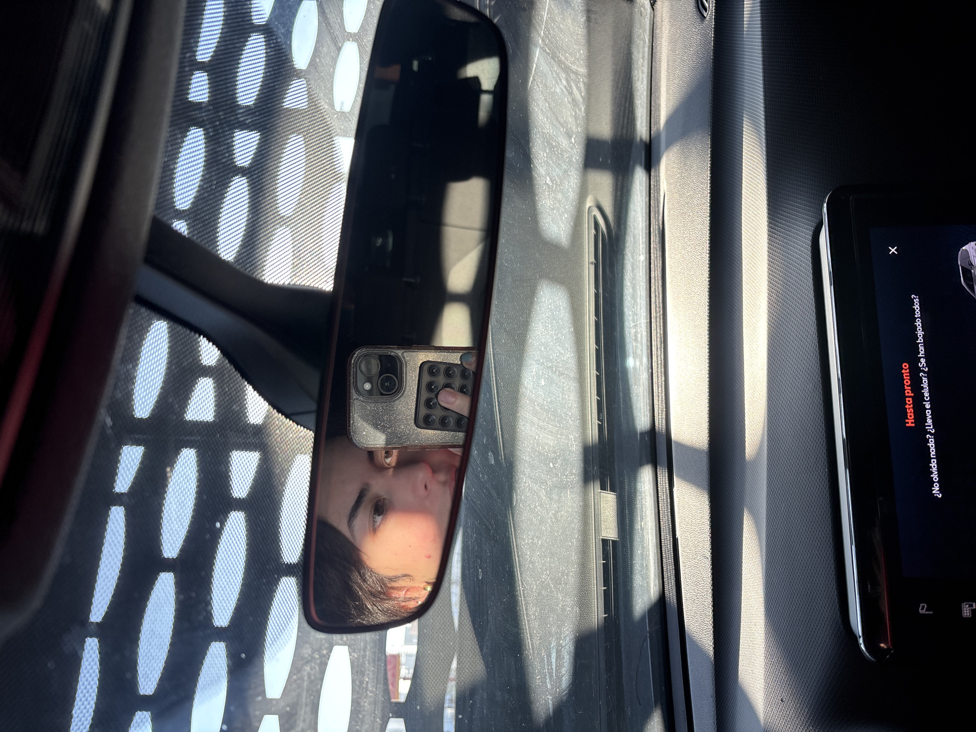 An image taken from inside a car in San Bernardino Tlaxcalancingo, Mexico, captures a reflection in the rearview mirror under bright daylight. The central feature is the car's rearview mirror, framed in a dark red/brown, reflecting a person's face, likely a young woman with dark hair and a neutral expression. She appears to be holding a smartphone with a glittery case and multiple camera lenses, which is also visible in the reflection, with her finger touching the back of the device.
To the right of the mirror, the car's interior paneling, possibly the dashboard, is visible, textured and illuminated by strong, directional sunlight that creates distinct shadows. Further right, a second smartphone screen displays text in Spanish. The visible text reads: "Hasta pronto" (See you soon), followed by "¿No olvida nada? ¿Lleva el celular? ¿Se han bajado todos?" (Don't forget anything? Do you have your cell phone? Have everyone gotten off?). An "X" button and a "P" icon are also visible on the screen.
Through the windshield, visible in the upper-left background, a patterned grid of light and shadow suggests a sunshade or textured roof panel outside the vehicle. The strong lighting indicates it is daytime. The scene conveys a moment of transition, perhaps before departure or after arrival, with the person interacting with their phone while another device prompts about leaving.