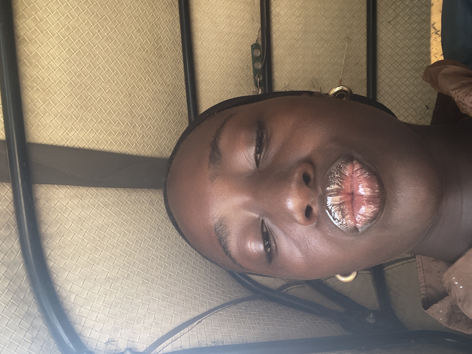 A close-up, rotated, and possibly inverted image features a dark-skinned individual, likely a female, with closed eyes and lips pursed. The lips have a glossy, reflective appearance. The background consists of a woven material, possibly a seat cover, with what appears to be a metal frame visible on the left side and potentially a piece of hardware or a clasp visible towards the top. The environment cannot be ascertained definitively based on the image alone. The location information of Galadimawa Village, Nigeria, is provided, but there are no visual cues within the photograph itself to confirm or contradict this assertion.