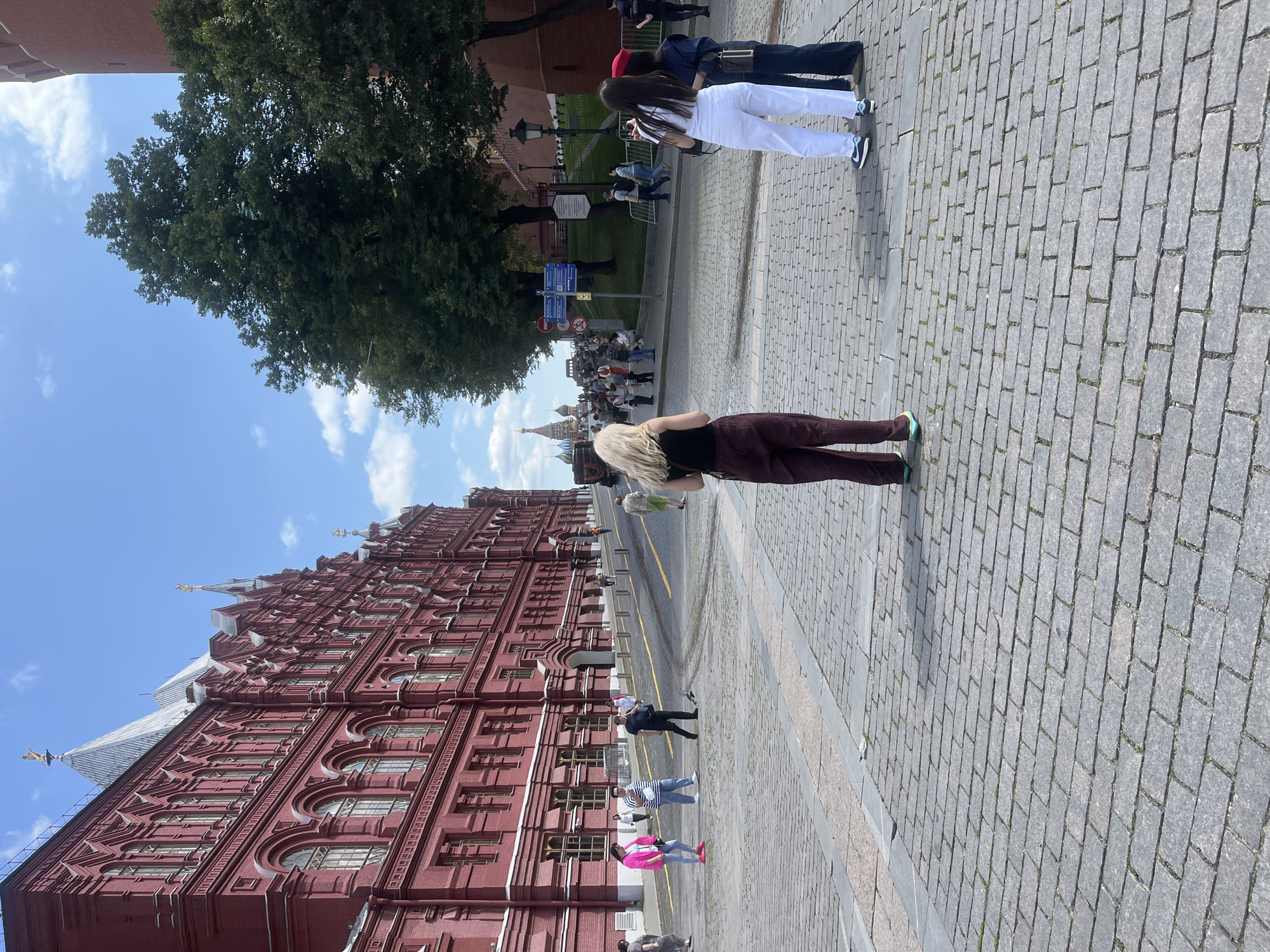 The image depicts a view of Red Square in Moscow, Russia, on a sunny day. In the foreground, three women are standing on a cobblestone pavement. The woman on the left has blonde hair and is wearing a black top and maroon pants. The woman in the middle is wearing a white top and white pants, with a red beret. The woman on the right is wearing a dark blue denim jacket and pants. Behind them, a large, ornate red building, the State Historical Museum, occupies the left side of the frame. Several people are walking in the background, and trees are visible on the right side of the square. In the distance, the distinctive onion domes of Saint Basil's Cathedral are visible. The sky is blue with scattered clouds.