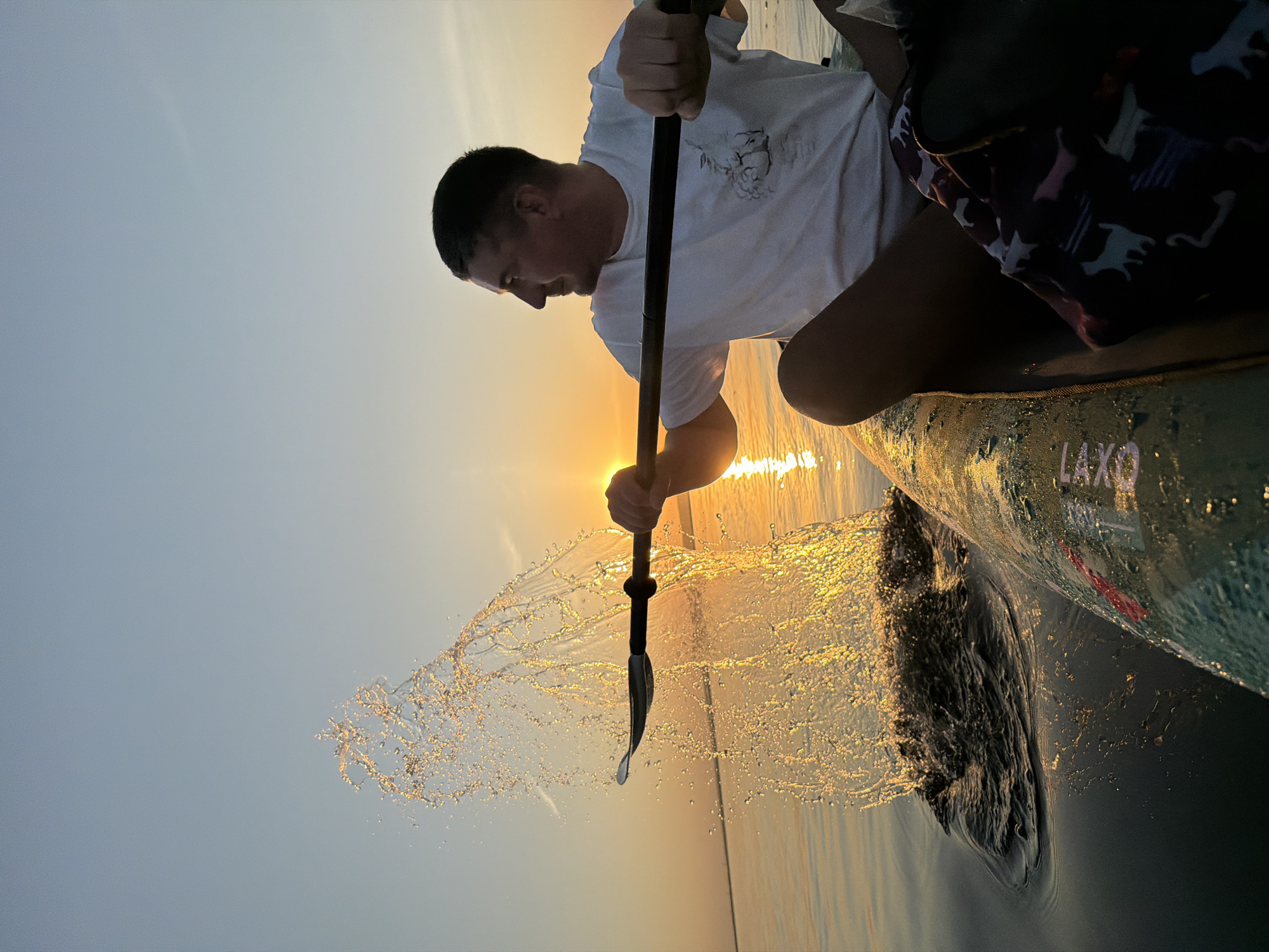 A man in a white t-shirt is paddling a paddleboard on the water during a sunset. Water splashes from the paddle. The location in the city of Chaikovsky, Russia, is not evident in the image.