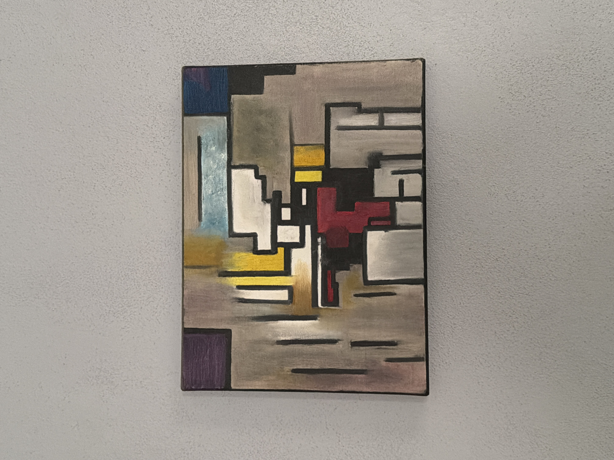A rectangular painting hangs on a white wall. The painting features a geometric, abstract design composed of squares and rectangles in various colors, including blue, purple, yellow, red, white, gray, and black. The background color is a mixture of taupe and gray. The painting's borders are black. The wall has a textured surface. The image does not contain any information regarding the city of Cuautla, Mexico.