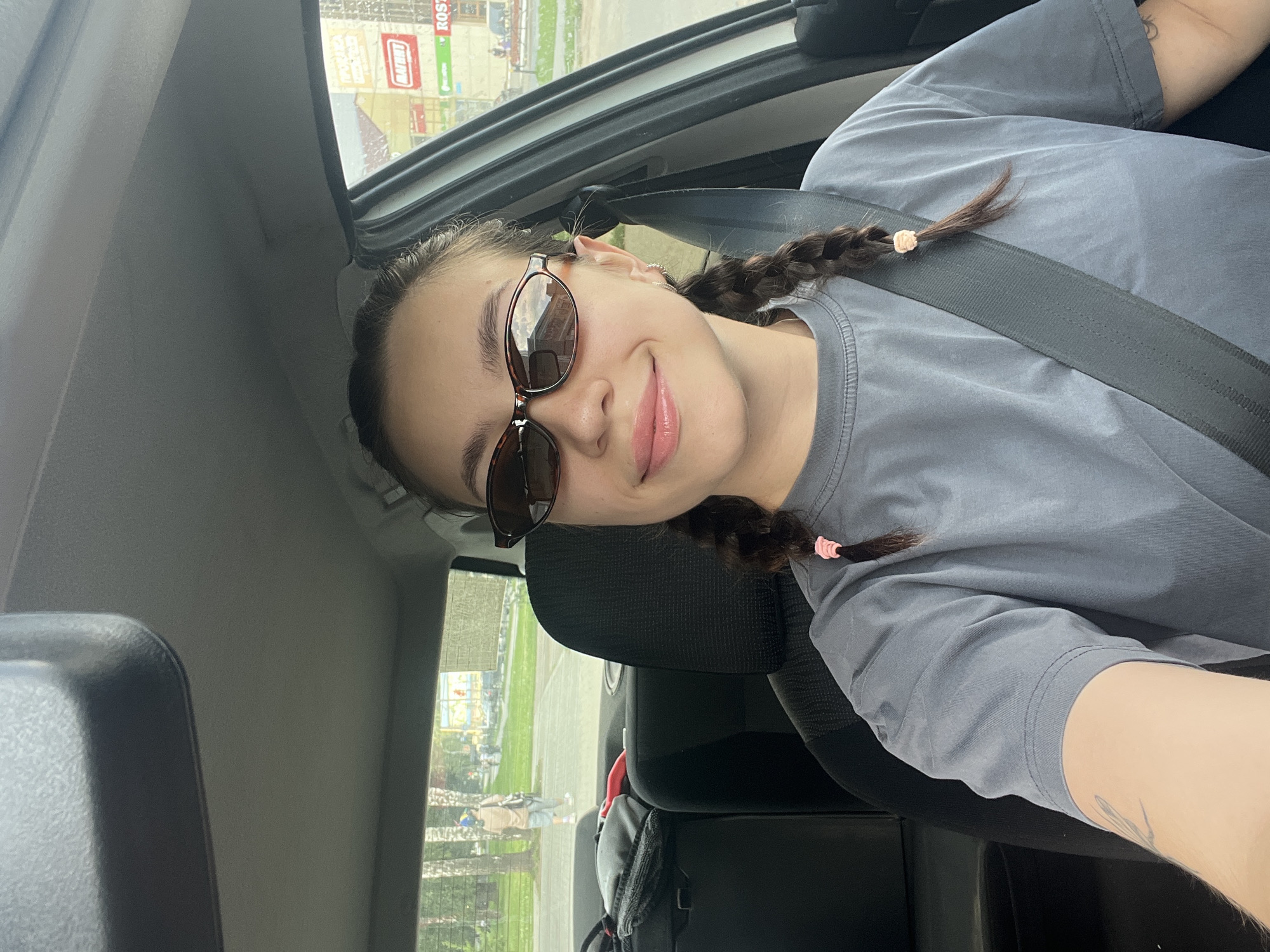 A young woman with brown hair in two braids, wearing sunglasses and a grey t-shirt, sits in the driver's seat of a car. She is smiling and looking at the camera. The car is parked on a street in Chaikovsky, Russia, with buildings and trees visible in the background. A seatbelt is fastened across her chest.