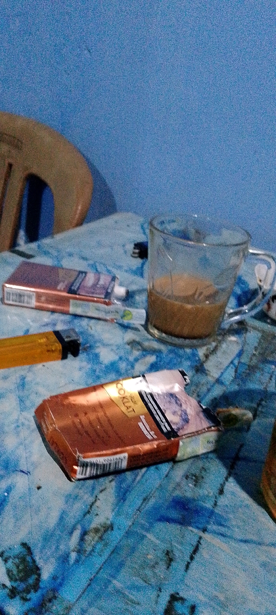 The image depicts a close-up, low-angle shot of a blue, textured surface, likely a table, with several objects on it. In the foreground, a partially opened cigarette pack with an orange-brown color and gold accents is visible. The text "COKLAT" is legible on the pack. To the left of this cigarette pack, a bright yellow lighter is positioned horizontally. Further back, another cigarette pack, also partially open and predominantly red and white, lies on the surface. Behind these items, a clear glass mug containing a light brown beverage, possibly coffee or tea, is situated. The background features a textured blue wall and the edge of a light brown chair. The overall scene suggests a casual, indoor setting. The image does not contain any elements that would allow for a specific location within Sukamukti, Indonesia to be identified.