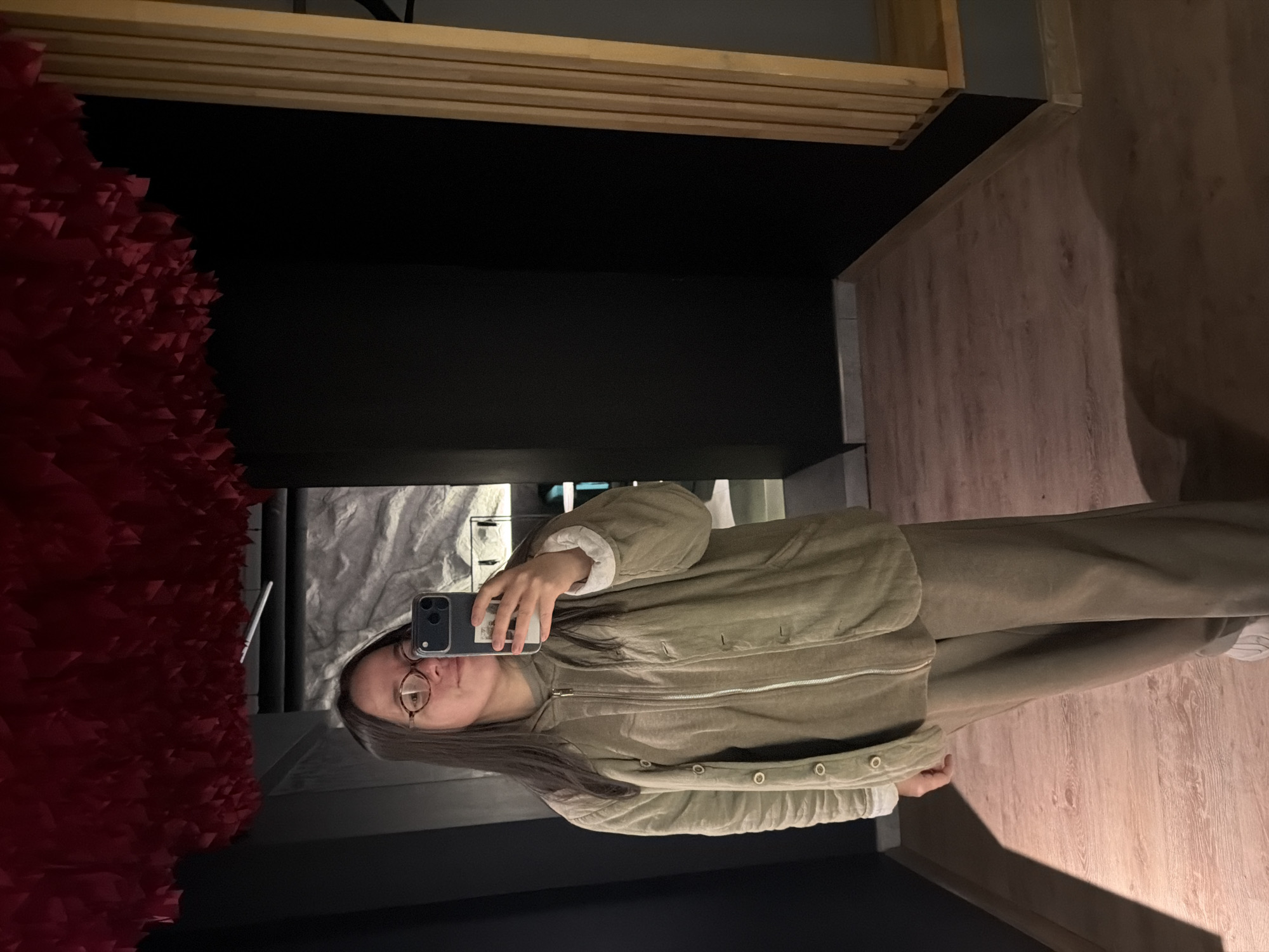 A young woman in glasses and tan clothing is taking a selfie in what appears to be a mirror. To her left is a dark wall with a red, textured surface. Behind her is a room with light-colored wood flooring and what seems to be a grey object. The photograph might have been taken in Chaikovsky, Russia.
