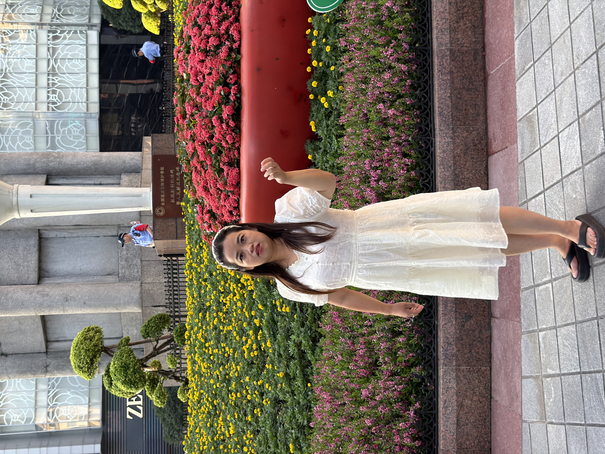 A woman with dark hair and a white dress stands in front of a large red planter filled with red flowers. Behind her, a dense patch of yellow flowers is visible, bordered by a black metal fence. In the background, a building with a grey, textured facade and large windows with white decorative patterns is partially visible. Two individuals in blue uniforms are standing near the building. The woman is facing forward with her right hand raised. The ground in front of her is paved with grey square tiles. The scene appears to be in an outdoor urban setting, likely in the Yuzhong District of China, given the presence of what appear to be security personnel and the architectural style.