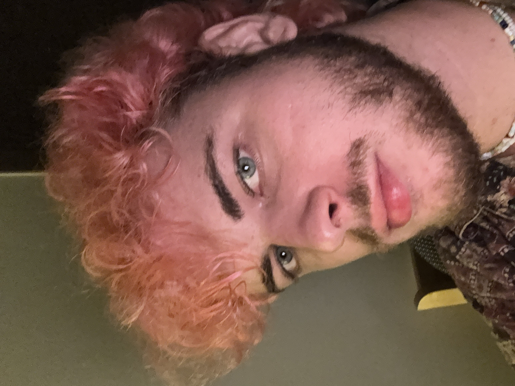 A close-up image shows a person with pink hair, light skin, a dark beard, and light blue eyes. The person is wearing a beaded bracelet and a patterned shirt. The background is a plain, solid color. The image is slightly tilted. There is no indication of Đà Nẵng, Vietnam, or any city location.