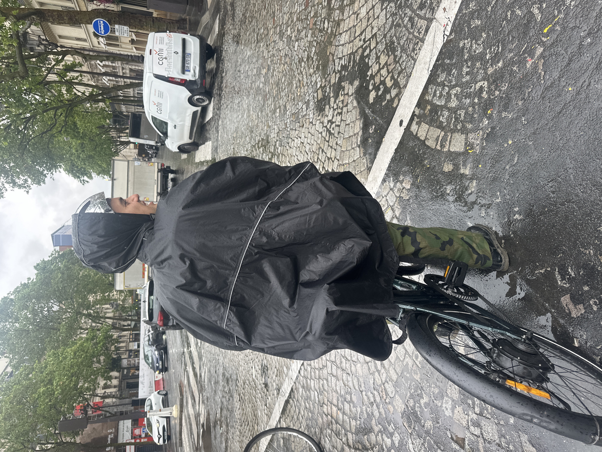 A person wearing a black rain poncho is riding a bicycle on a wet cobblestone street. The person is also wearing a helmet and camouflage pants with black boots. The street is lined with buildings and trees. In the background, a white van with the word "CGMI" is parked. Other vehicles are visible in the distance. The street appears to be in Paris, France. The presence of CGMI van and the architecture indicates that.
