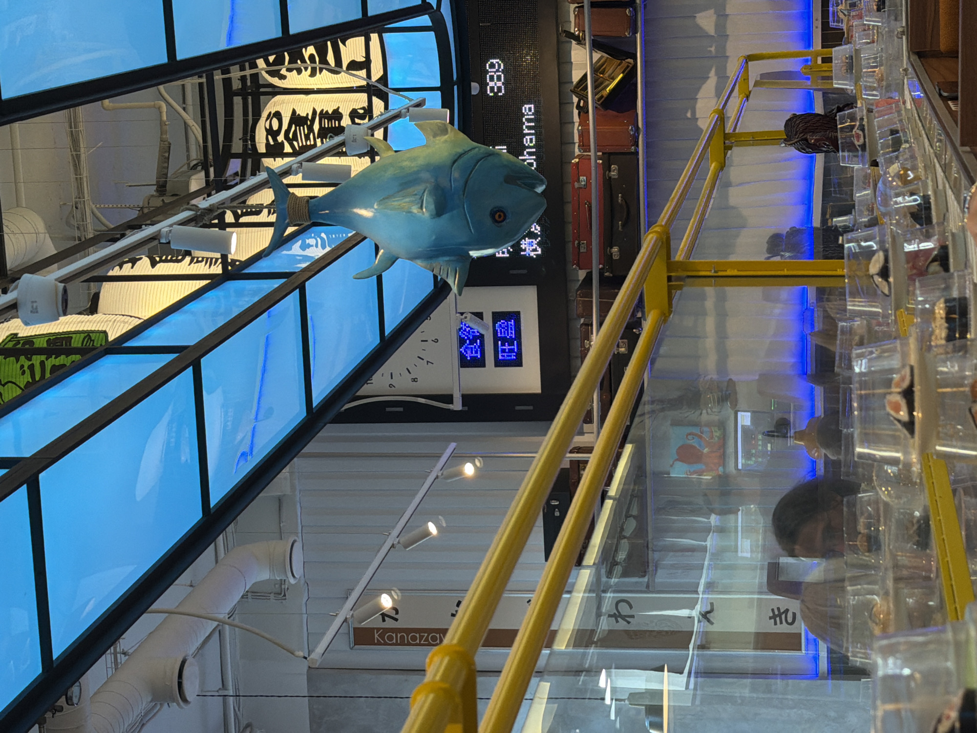 This image depicts an interior scene, likely a restaurant, with a conveyor belt system for serving food. A large, turquoise fish decoration is suspended from the ceiling. In the background, a digital display shows "Yokohama" and the number "389." The ceiling features a bright blue illuminated panel. The conveyor belt is lined with numerous small, covered plates of food, likely sushi. Reflections in the transparent partitions reveal people and restaurant decor. The overall ambiance suggests a modern, potentially Japanese-themed establishment. The location provided, Saint Petersburg, Russia, is a detail that cannot be independently verified from the image alone.