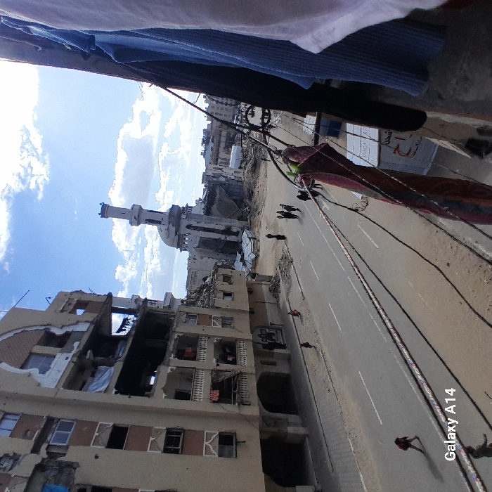 A war-torn street is framed by damaged buildings, one severely impacted, with a mosque standing amidst the devastation. Power lines crisscross the scene, and figures walk the empty road. Laundry hangs overhead, adding an element of ordinary life to the harsh backdrop. The sky is a mix of blue and clouds. The photograph, captured with a "Galaxy A14", shows a scene of urban destruction and resilience.