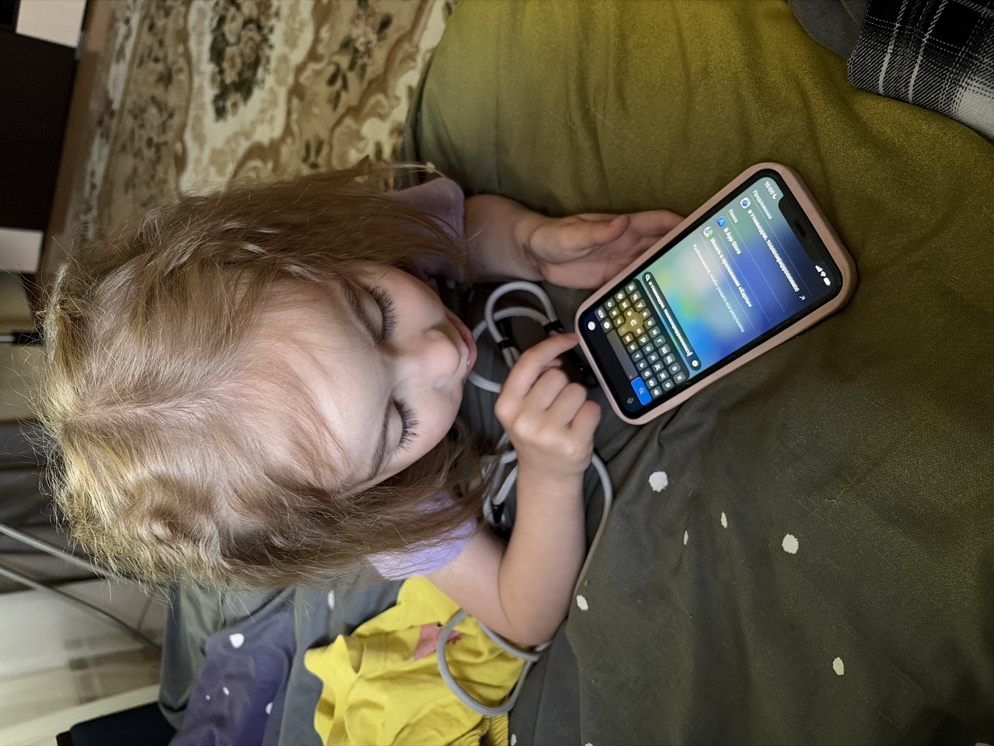 A young blonde-haired child is lying down, looking at a smartphone. The child is wearing a purple top and a yellow shirt. A cord is wrapped around the child's arm and the phone. The child's eyes are closed, and they appear to be concentrating or perhaps dozing. The phone is held in the child's left hand, with the right hand reaching towards the screen. The phone displays a search interface with text in Russian. Visible text on the screen includes "Предложения" (Suggestions), "Поиск" (Search), "App Store", and "Искать в приложении «Карты»" (Search in the «Maps» app). The time displayed on the phone is 15:00. The background suggests an indoor setting, possibly a bed or couch, with a patterned rug or blanket visible. The child is lying on a dark green fabric with white polka dots. The lighting suggests it is daytime, but no external weather cues are available. The overall scene conveys a sense of quiet focus or relaxation as the child interacts with the device.