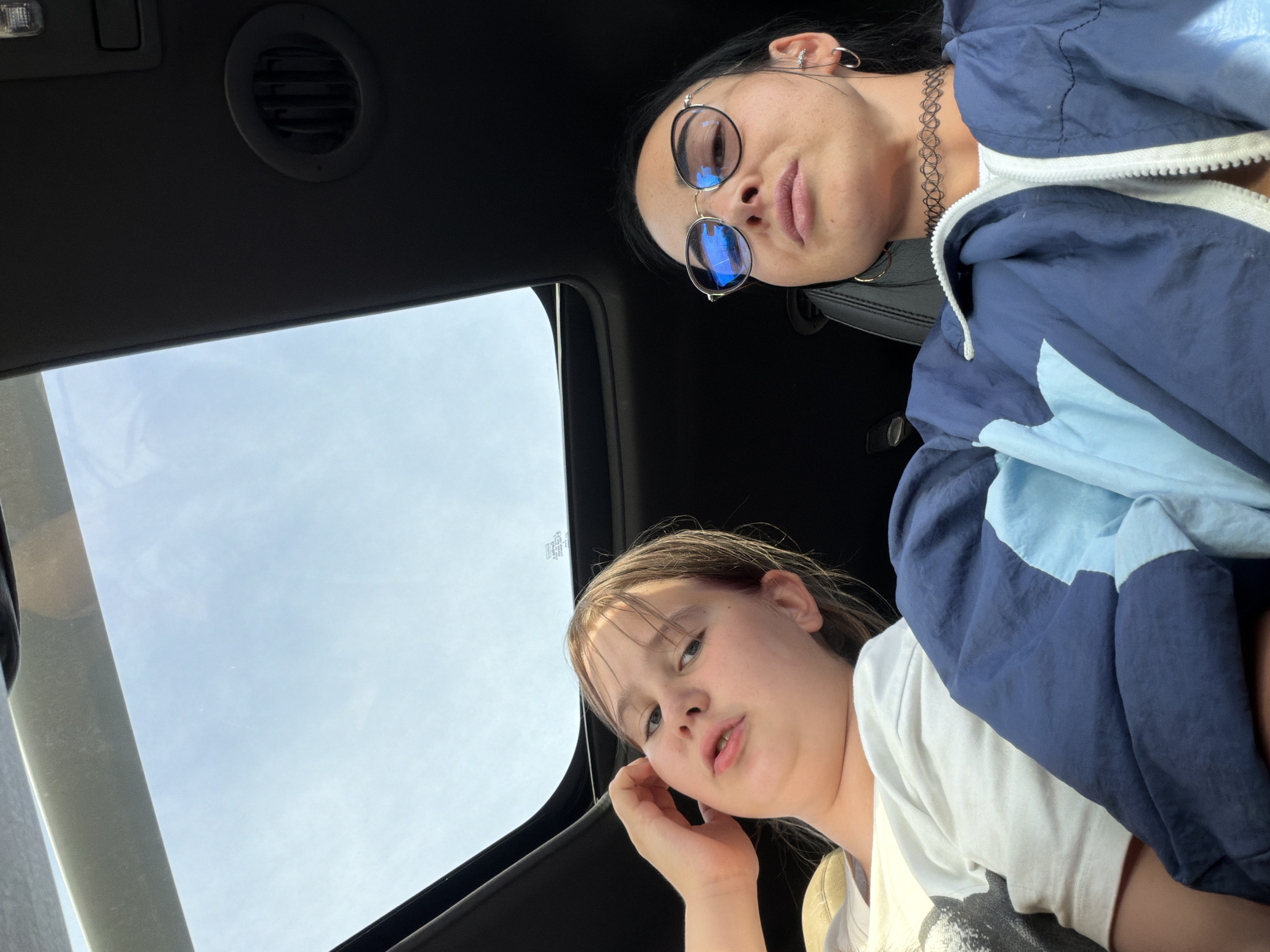 Two young females are inside a vehicle, looking up at the camera through the sunroof. The subject in the foreground is a girl with blonde hair and blue eyes, wearing a white t-shirt. She has her right hand raised to her cheek, and her mouth is slightly open. Behind her, a young woman with dark hair wears round sunglasses and a choker necklace. She is looking down at the camera and is dressed in a blue and light blue jacket. The scene appears to be outdoors during the day, with a clear sky visible through the sunroof. The interior of the car is dark, with a vent visible on the ceiling. The image is a selfie taken from a low angle.