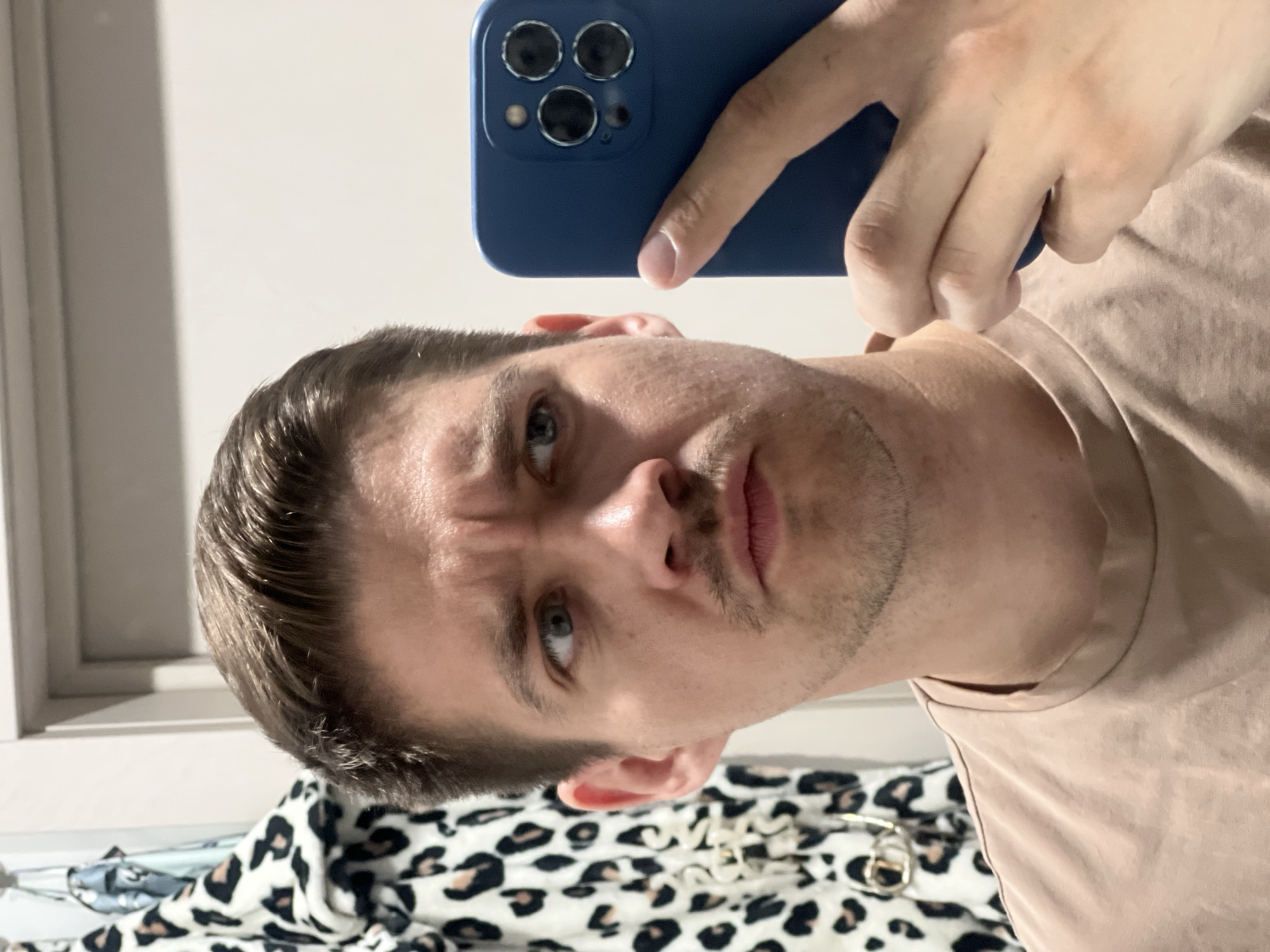 A man with short brown hair and blue eyes is taking a selfie using a blue smartphone. He is wearing a beige t-shirt. A leopard print blanket is visible in the lower portion of the image. The location is not discernible from the image.