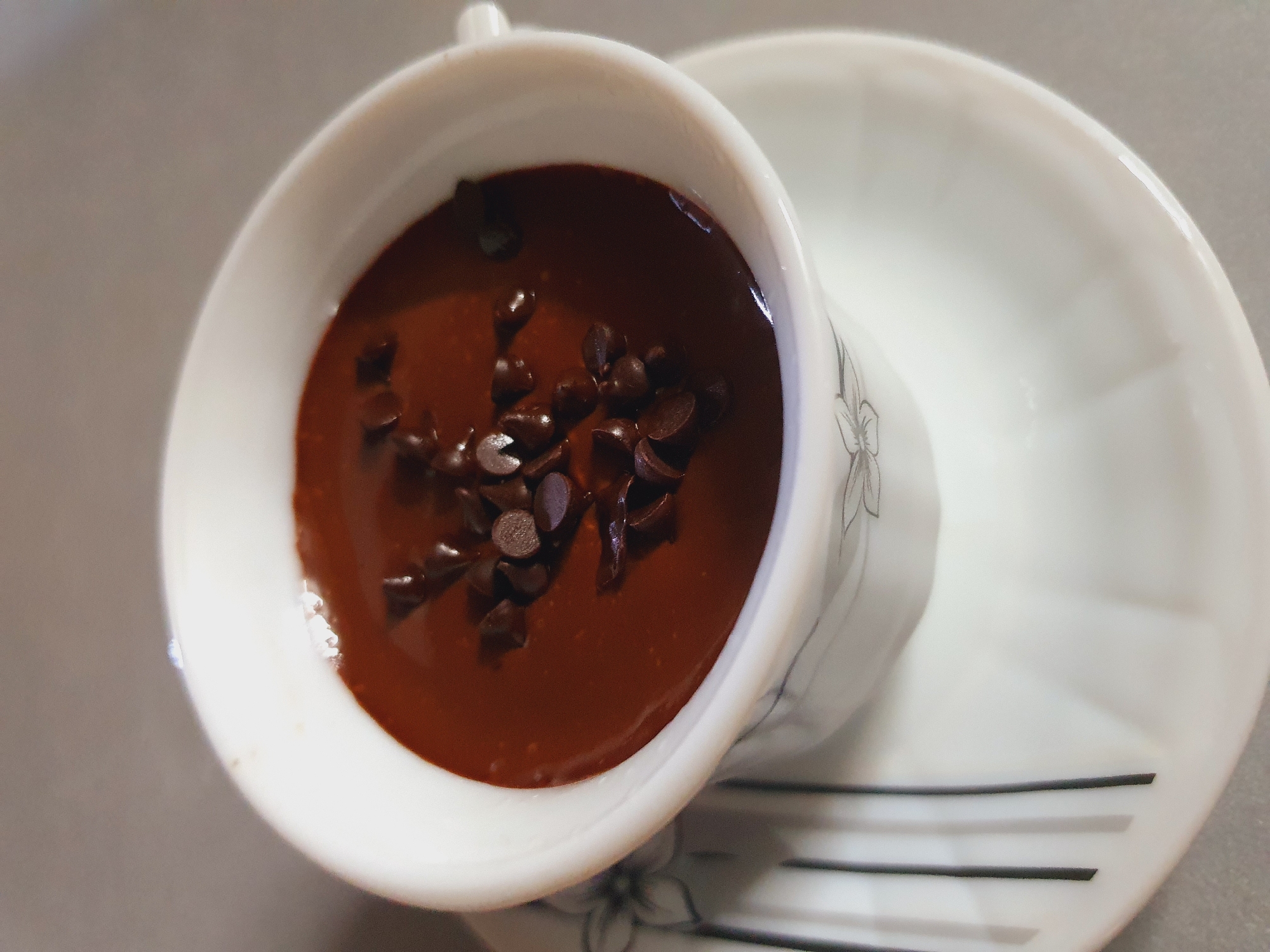 A close-up, overhead shot shows a white teacup filled with dark chocolate pudding, topped with a scattering of chocolate chips. The teacup has a delicate floral pattern around its rim and sits on a matching saucer. The saucer also features the same floral design and has several dark vertical stripes on its left side. The background is a neutral, light gray surface, suggesting an indoor setting. There is no indication of the city of Shiraz, Iran.