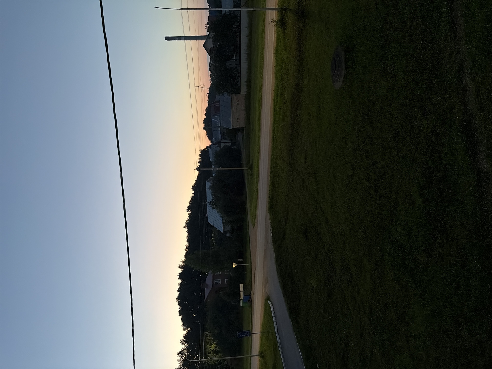 This media file shows a rural scene in Birsk, Russia, at dusk. The sky transitions from a clear blue to a soft orange and yellow hue near the horizon, indicating the setting sun. A thin, dark power line stretches diagonally across the frame from the lower left corner, suggesting the presence of electricity infrastructure in the area.
The landscape features a dirt road winding through a grassy area. On one side of the road, several houses and buildings are visible, nestled amongst dark green trees. Some of these structures have pitched roofs, typical of residential buildings. A tall industrial chimney stands prominently in the background, hinting at nearby industrial activity. A few streetlights are also present, adding to the twilight ambiance.
On the other side of the road, the terrain is covered in dense, dark green grass, with a circular object that appears to be a manhole cover or grate embedded in it. The overall atmosphere is peaceful and quiet, typical of a rural evening. There is no visible text in the image.