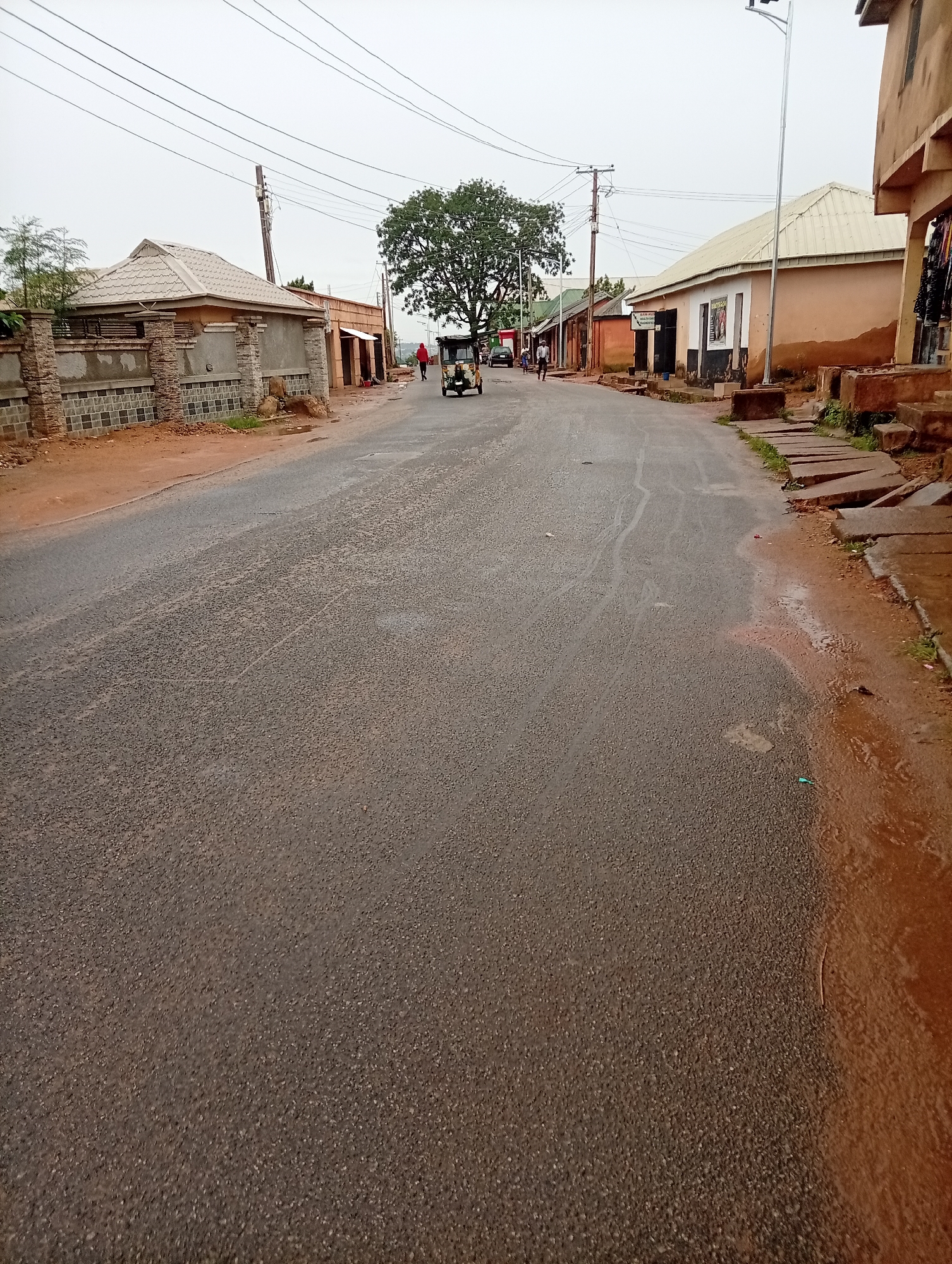 A street in Gura Top, Nigeria is viewed lengthwise, receding into the distance. The street is asphalt and appears damp. Along either side of the street are buildings constructed primarily of ochre-colored material. The buildings are single-story structures with corrugated metal roofs. A large tree is visible at the center of the street, providing shade. Several people are walking along the street, and a three-wheeled vehicle is also present. Power lines stretch overhead, and a street light is visible on the right side of the frame. The scene conveys a sense of everyday life in a residential or commercial area of Gura Top.