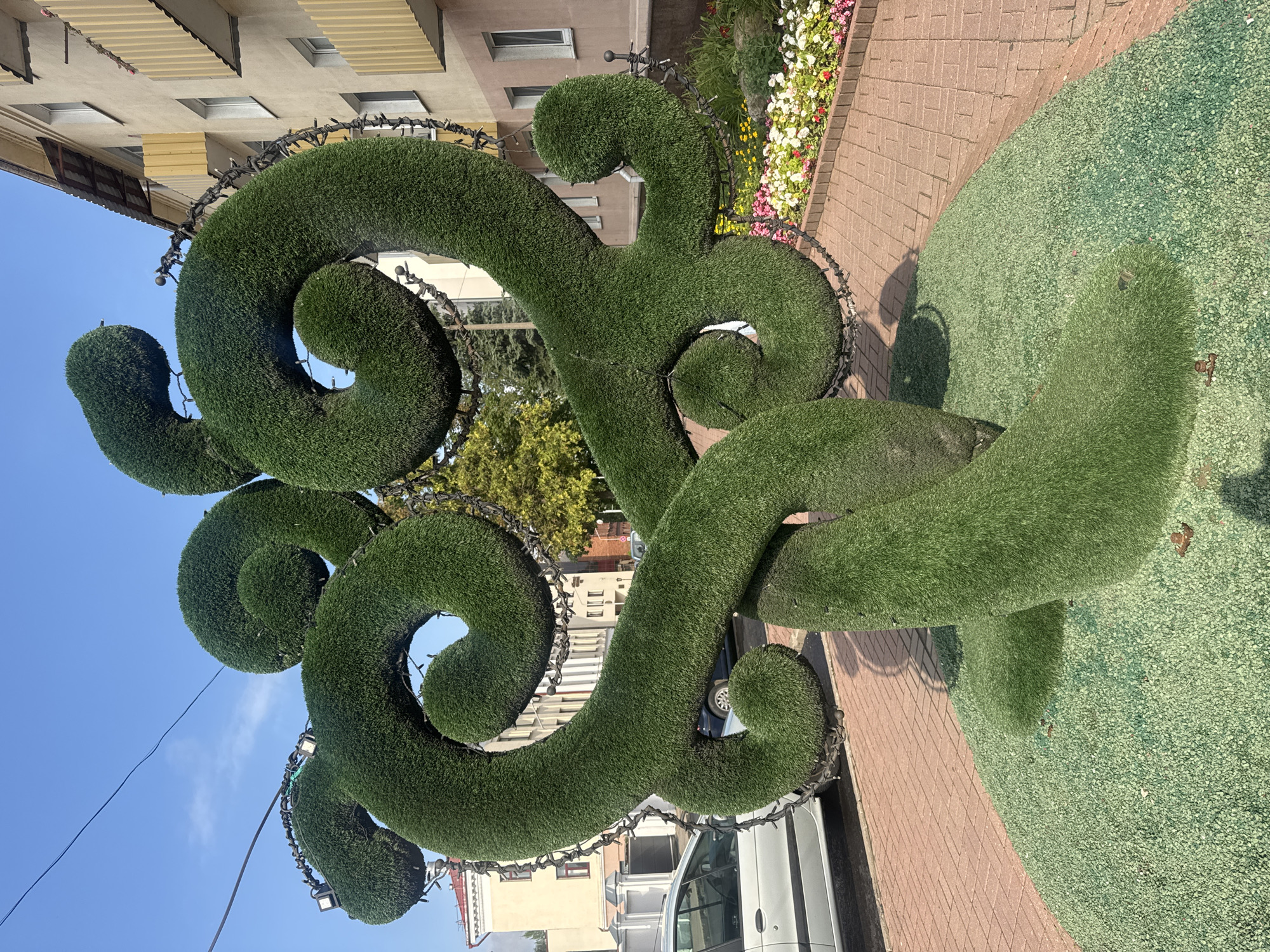 The image shows a large, spiral-shaped topiary sculpture covered in artificial grass, located outdoors in a city. The sculpture is green and has a rough texture. It is decorated with strings of small lights and some spotlights attached to its structure. The sculpture sits on a base covered in green gravel. In the background, there is a building with a yellow facade, balconies with yellow railings, and windows. There are also some trees and a flowerbed with colorful flowers. A brick sidewalk is visible in front of the flowerbed and extends towards the sculpture. A silver car is parked on the left side of the image. The sky is blue with some wispy clouds, and electrical wires are visible overhead. The lighting suggests it is daytime, possibly late morning or early afternoon, casting shadows from the sculpture.