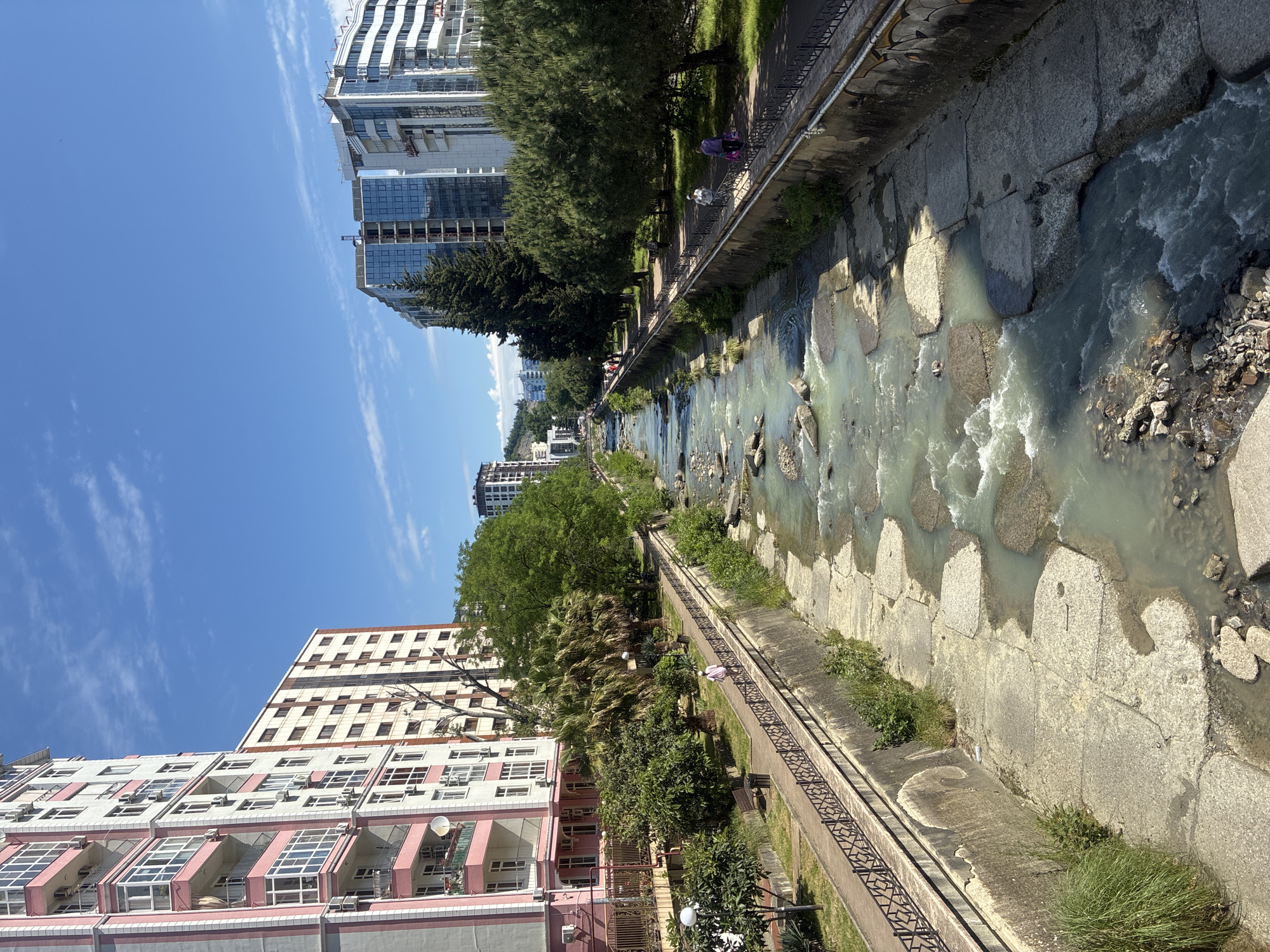 The image captures an urban river scene in Sochi, Russia, under a bright, clear blue sky with a few wispy clouds, suggesting a sunny day, likely mid-day or early afternoon given the distinct shadows. The river, with greenish-blue, turbulent water and exposed concrete and rock formations, flows through the center of the frame. Its banks are lined with concrete revetments, some with grass and small bushes growing.
Along the river, a paved pedestrian pathway with a black metal fence provides a recreational space. Several people are visible on this path: two individuals, one with a purple head covering and another in a white top, are walking in the mid-ground, while another person is further down near a pink building. Lush green vegetation, including coniferous trees, palm trees, and deciduous trees, lines both sides of the river, creating a verdant corridor within the city.
Flanking the river are various urban buildings. On the left, a multi-story residential building with a distinctive pink and white facade features numerous balconies and visible air conditioning units. Further back, another tall building with a grid pattern of windows and a lighter facade is visible. On the right side of the river, modern high-rise buildings, some with glass facades and balconies, dominate the skyline. One building appears to be under construction, with visible cranes and scaffolding. The architecture showcases a mix of contemporary and slightly older styles. No discernible text is visible in the image.