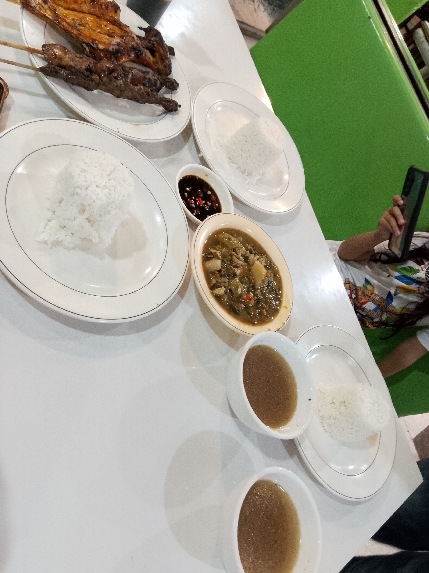 A table setting at a restaurant in Davao City, Philippines, features a meal consisting of grilled chicken skewers, rice, a stew-like dish, and two bowls of soup. The grilled chicken, appearing to be chicken parts on skewers, is placed on a white oval plate. Adjacent to it, another white plate holds a mound of white rice. A small bowl with a dark sauce, possibly soy sauce with chili, is also visible. In the center of the table, a larger oval bowl contains a stew with vegetables and possibly meat. Two smaller white bowls are filled with a light brown liquid, likely soup. To the right, a person, partially visible, is holding a smartphone, with their arm and shoulder showing. They are wearing a white t-shirt with a colorful print. The table is white, and the background includes a bright green wall. The lighting suggests an indoor setting, possibly during the day or with artificial lighting.
