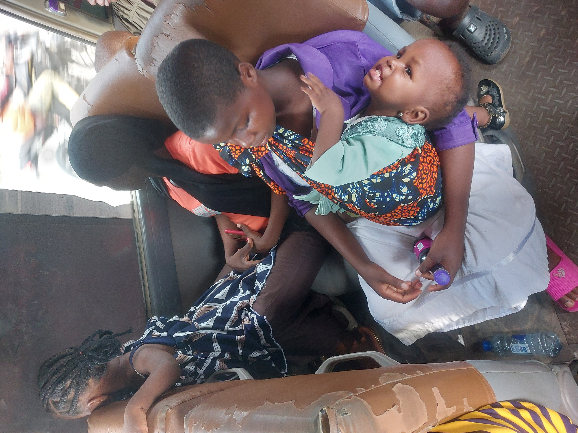 A medium shot depicts a crowded interior scene, possibly a public transport vehicle in Dar es Salaam, Tanzania, given the presence of African individuals and environmental clues.
The focal point is a group of four individuals. On the left, a young girl with braided hair rests on the seat. Next to her is an individual holding a mobile phone, wearing a patterned top, brown trousers, and a long black braid. Adjacent to this individual is a second individual, wearing a purple top and patterned orange and blue shirt, holding a baby on their lap. The baby is dressed in clothing of similar patterned orange and blue fabric with a light green top and a small silver earring. They are holding a small purple bottle.
A light-colored, possibly white cloth covers the lap of the individual with the baby, and a water bottle rests near their feet, clad in pink sandals. The vehicle floor has a dark, metal texture.
The environment is crowded and typical of public transport, with worn-out seat covers and a general lack of space. Other partially visible passengers are seen in the periphery.